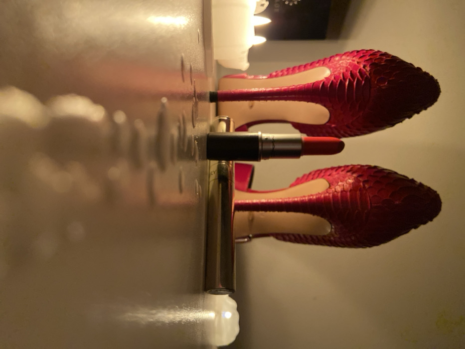 A photograph depicts red snakeskin heels and makeup arranged on a shiny surface, possibly a mirrored vanity. The heels are reflected below, creating symmetry. A black and silver lipstick tube lies horizontally between the shoes, and a gold tube stands vertically near the bottom. A blurry white object sits at the edge of the frame near the gold tube. The lighting casts soft shadows, enhancing the reflective surface and the textures of the shoes.