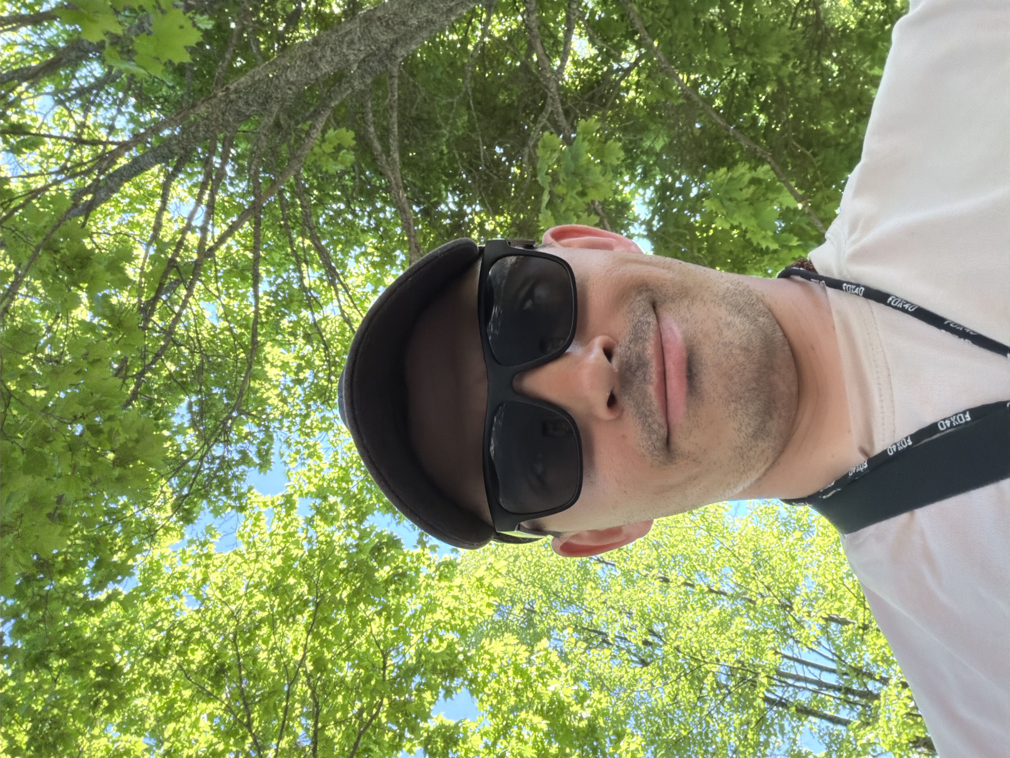 A man wearing a black flat cap and sunglasses is facing the camera. He also wears a white shirt and a black lanyard with "FOX40" printed on it. The camera angle is looking upwards at the man, with green trees and a clear blue sky visible in the background. The location is outdoors, with the trees and sky suggesting it could be a park or wooded area. The image is likely taken in Oreshniki, Russia, due to context. The man appears to be taking a selfie.