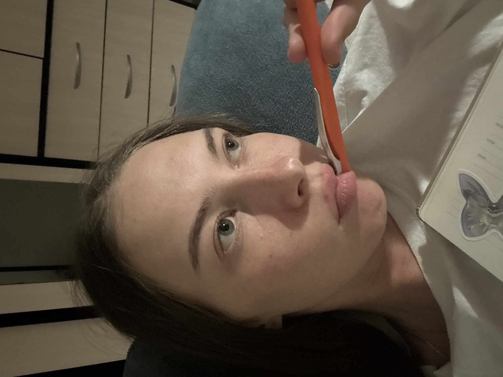 The image shows a close-up of a young woman's face, with her head slightly tilted. She is holding an orange pen to her lips, seemingly in thought. Her eyes are open and looking to the right. She is wearing a white t-shirt, and a necklace is visible. To the right of the woman, a blue textured surface, possibly a blanket or cushion, is visible. In the background, a dresser with three drawers is seen. The lighting suggests an indoor setting, likely a bedroom. The woman appears to be in a relaxed or pensive mood. There is no indication of the city of Perm, Russia, within the image itself.