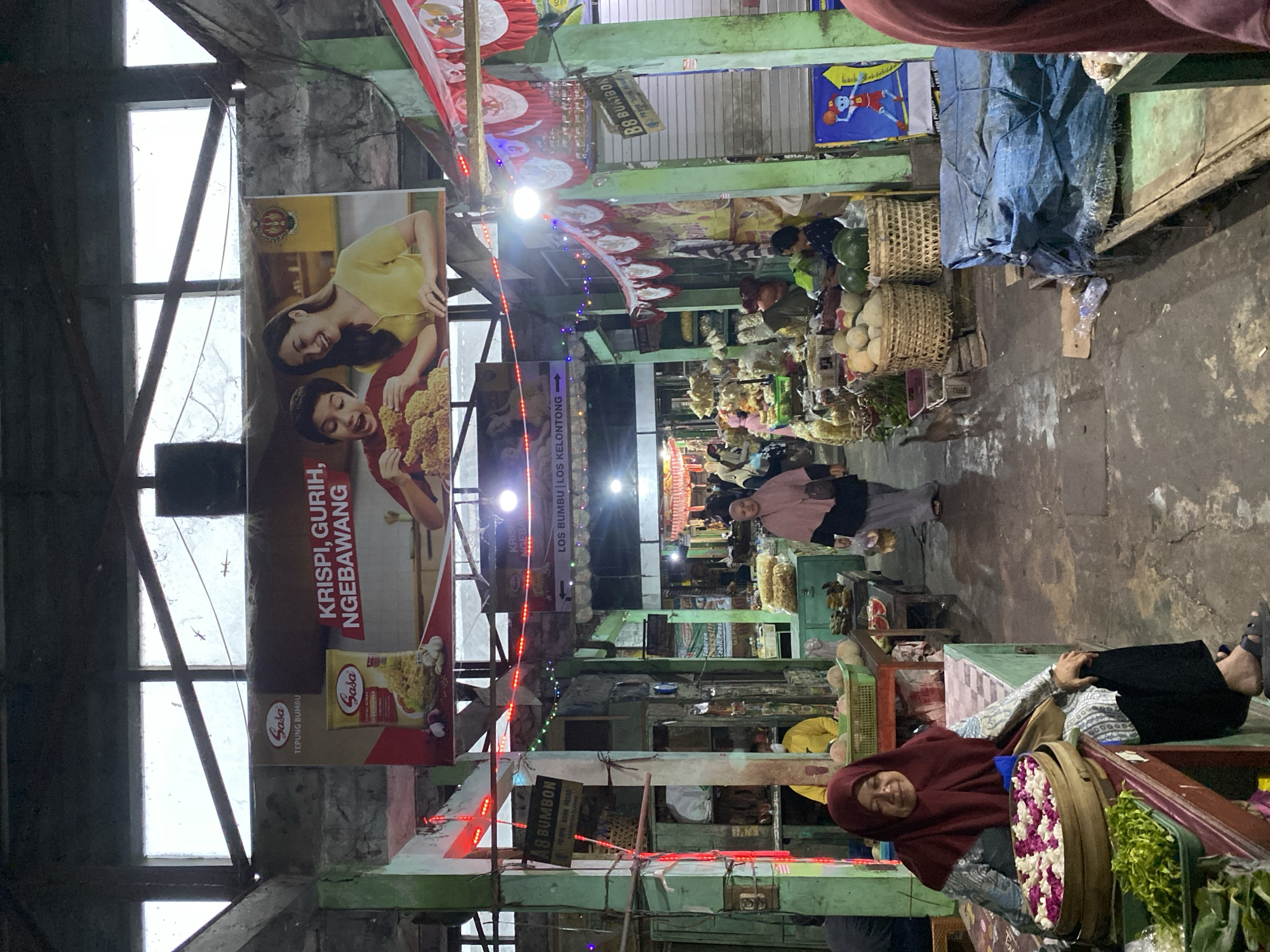 The image depicts an indoor market in Ambarketawang, Indonesia. The market has a high ceiling with large, translucent panels that allow diffused natural light to enter. A large banner advertising "Sasa Tepung Bumbu" is prominently displayed at the top, featuring a child and a woman eating fried food. The market stalls are organized along a central walkway, with vendors selling various goods including produce, spices, and other household items.
In the foreground, a woman wearing a maroon hijab and patterned clothing sits behind a stall laden with flower petals in a wooden container and green leafy vegetables. She is looking towards the camera with a slight smile. Further into the market, other vendors are seen attending to their stalls, and customers are browsing. The stalls are constructed from wood and metal, with some featuring colorful decorations and signage. There are also strings of festive lights and red and white pennants hanging from the ceiling, suggesting a celebratory or market day atmosphere. The ground appears to be made of concrete and shows signs of wear and tear. The overall impression is of a bustling local market scene.