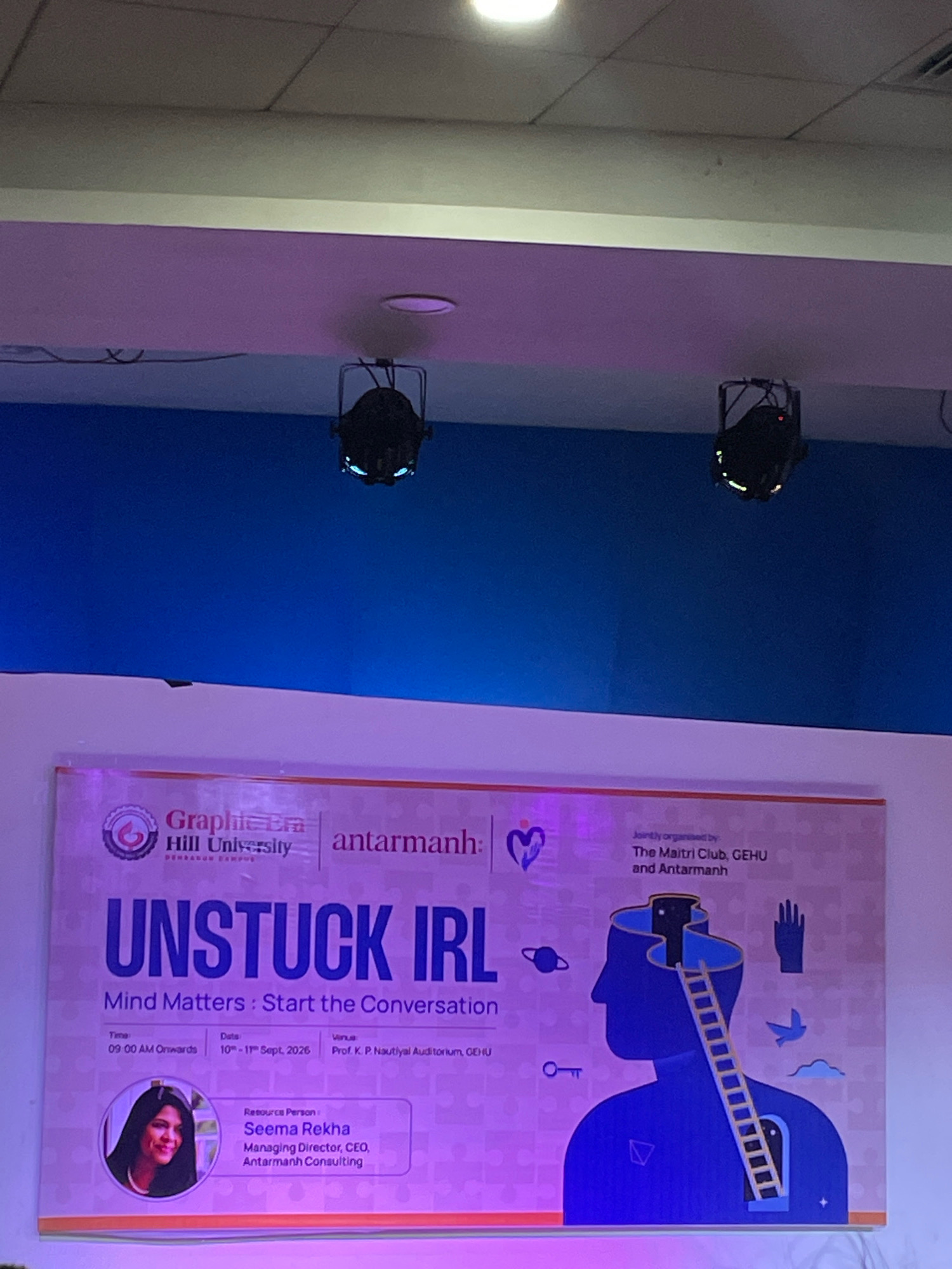 The image shows a banner for an event titled "UNSTUCK IRL: Mind Matters: Start the Conversation." The banner is purple and pink with a puzzle piece border. It features graphics of a silhouette of a head with a ladder leading into it, a hand, a bird, clouds, a planet, a key, and a doorway. The banner also includes the logos of "Graphic Era Hill University, Dehradun Campus" and "antarmanh."
The text on the banner provides event details:
* **Time:** 09:00 AM Onwards
* **Date:** 10th - 11th Sept, 2026
* **Venue:** Prof. K. P. Nautiyal Auditorium, GEHU
* **Resource Person:** Seema Rekha, Managing Director, CEO, Antarmanh Consulting. Her portrait is visible in a circular inset on the left side of the banner.
* **Jointly organised by:** The Maitri Club, GEHU and Antarmanh.
The banner is displayed in what appears to be an auditorium or large hall. Above the banner, the ceiling is visible, with fluorescent lighting and two hanging stage lights. The overall lighting suggests an indoor event setting. The purple and pink lighting on the banner implies stage or ambient lighting. The image is taken from a slightly low angle, looking up at the banner and the stage setup.