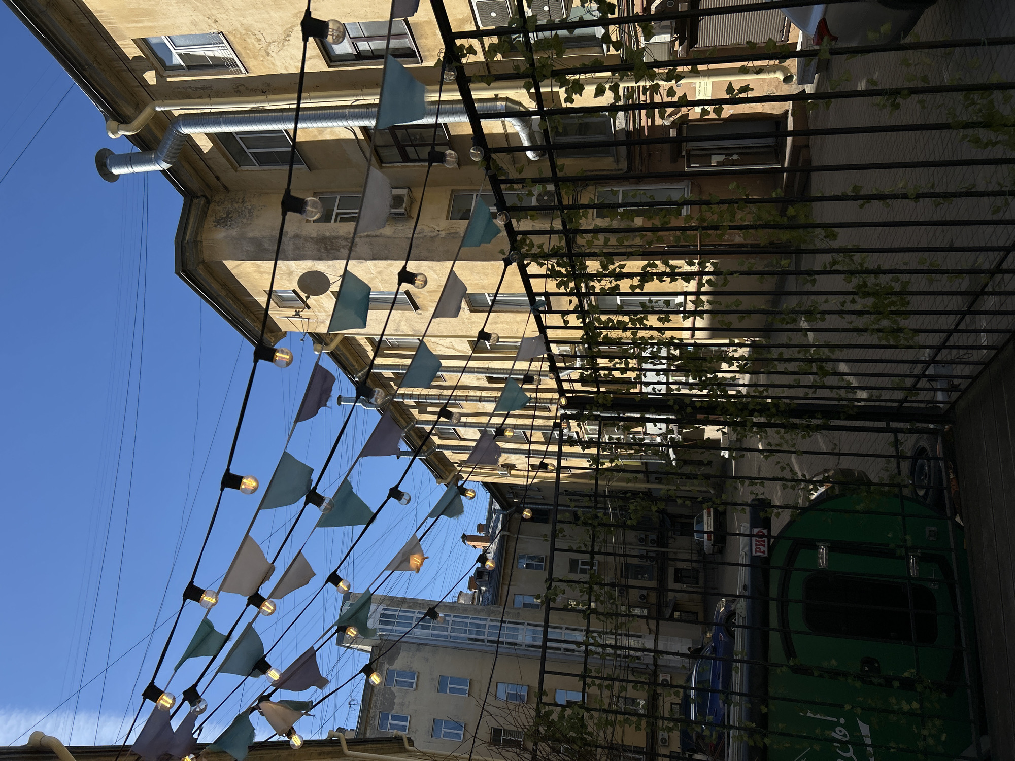 The image captures an upward view into a narrow urban space, likely a courtyard or passage in Saint Petersburg, Russia. Old, ochre-colored multi-story buildings dominate the background, featuring numerous windows, some with security bars, and exposed ventilation pipes and air conditioning units. The architectural style suggests an older European city.
Strung across the open space overhead are decorative elements: strings of lights with bare, warm-toned Edison bulbs and triangular bunting flags in shades of light blue, white, and light purple, creating a festive ambiance. A dark metal trellis, adorned with artificial green ivy, extends into the frame from the right, partially obscuring the view and casting strong, defined shadows onto the buildings and ground below, indicating bright, clear sunlight. Above the buildings, a vibrant, clear blue sky is visible, crisscrossed by thin electrical wires.
No people are visible, and there is no explicit activity taking place, though the decorations imply a welcoming or celebratory atmosphere. Through the trellis on the lower right, parts of a dark green vehicle, possibly a truck or bus, and a blue vehicle are partially visible. On the green vehicle, some indistinct white Cyrillic text is visible, including what appears to be "об!" or "сб!" followed by an exclamation mark. On a dark pole or structure next to the green vehicle, the Cyrillic letters "Рю" are discernible. The time of day is clearly daytime, sunny with strong shadows.