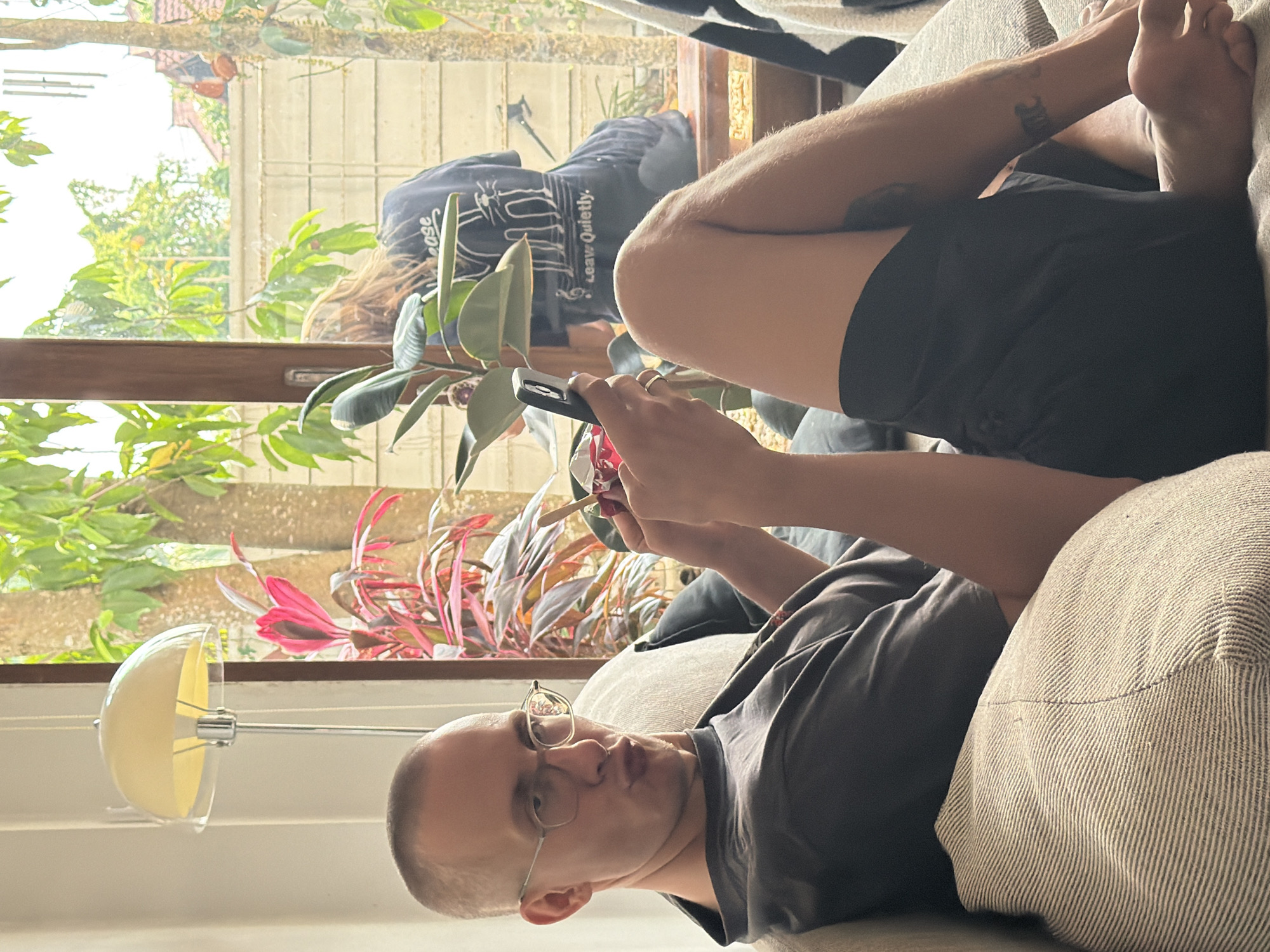 A person with glasses is reclining on a couch, holding a mobile phone. The person has a shaved head and wears a gray t-shirt. They are propped up against a striped cushion. Another person wearing a dark-colored shirt and light-colored hair is visible in the background, seated near a window. A plant with red and green leaves sits on the windowsill. Outside the window, trees and buildings are visible. The setting appears to be indoors. The location is inferred to be Chang Phueak, Thailand.