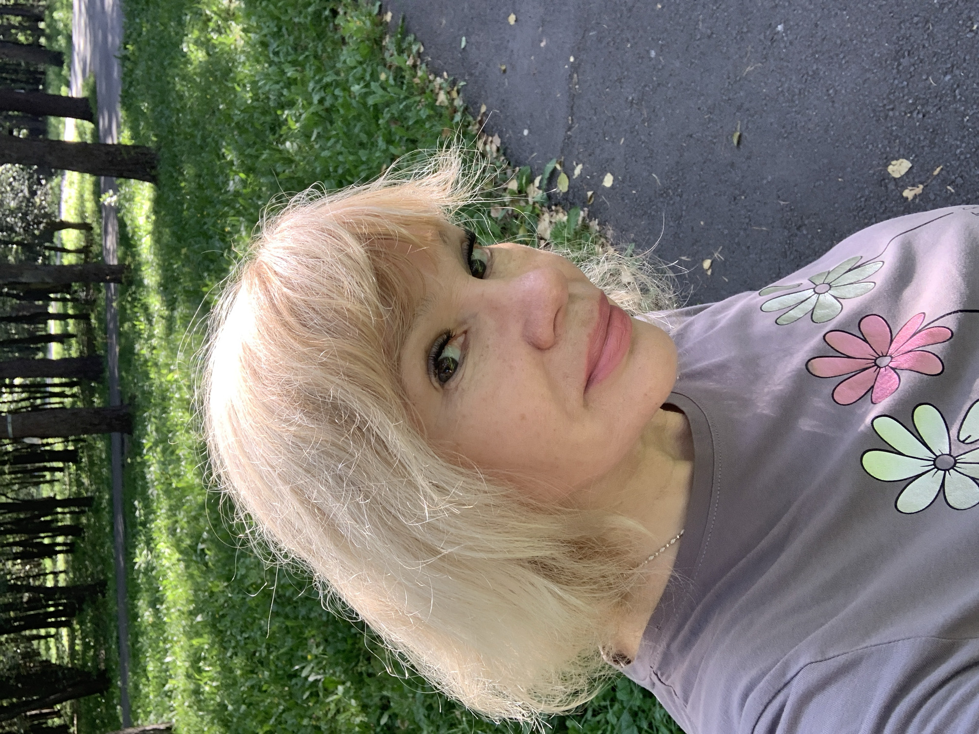 A middle-aged Caucasian woman with blonde hair and fair skin is captured in a close-up, outdoor selfie. She is wearing a grey t-shirt with a floral print featuring two flowers, one pink and one green. The woman has on makeup, including pink lipstick and mascara. Her hair is slightly disheveled, with wisps framing her face. The background suggests a park setting, with lush green grass, trees, and a paved path. The lighting indicates it is daytime, likely late morning or early afternoon. The image does not provide any specific information to determine its location within the city of Prokopyevsk, Russia.