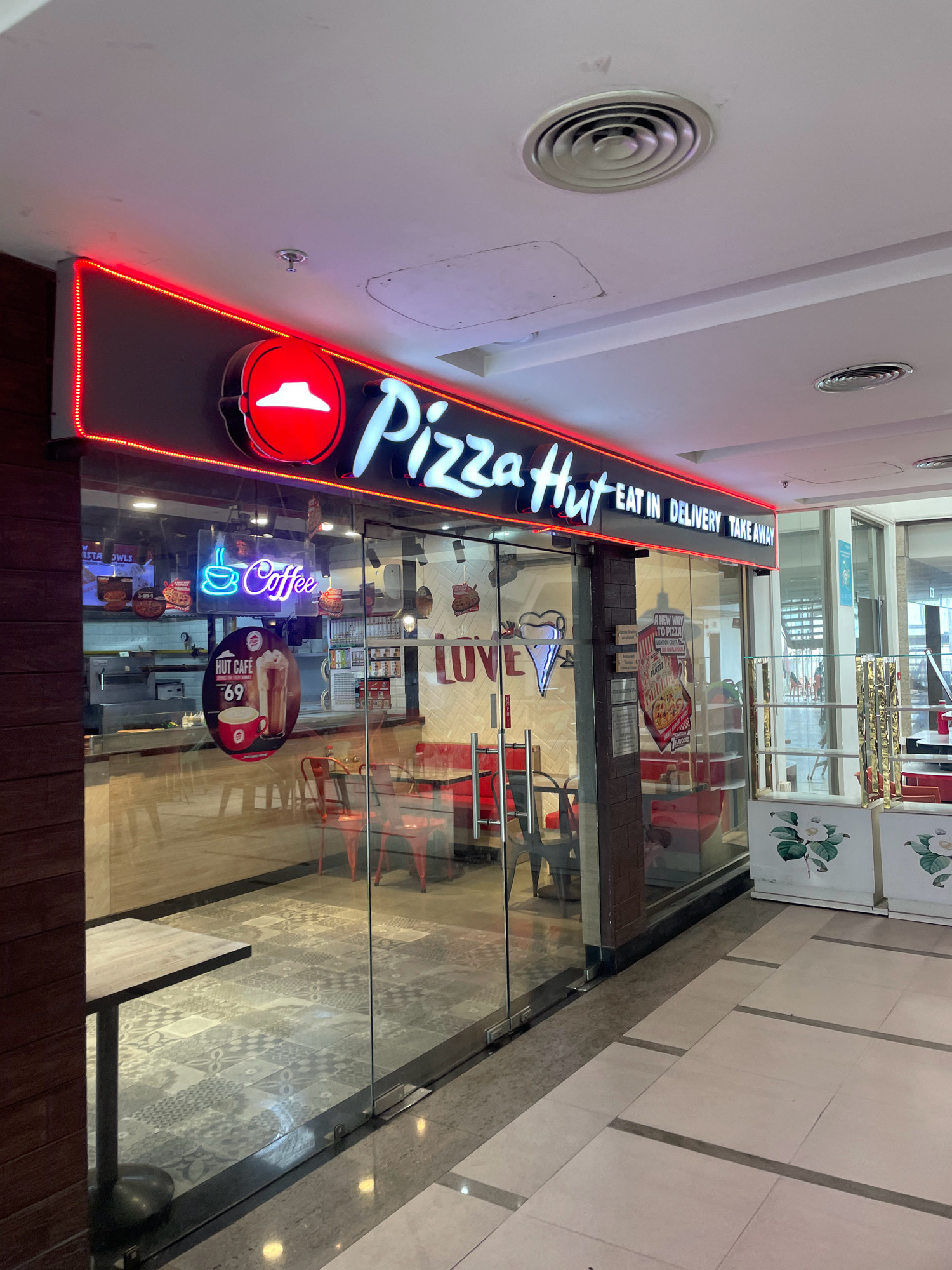 The image shows the exterior of a Pizza Hut restaurant located in a shopping mall in Dharamshala, India. The storefront is made of glass, and the prominent signage reads "Pizza Hut" in large white lettering with a red outline. The words "EAT IN DELIVERY TAKE AWAY" are also visible on the signage. Red neon lights outline the top of the storefront. Inside the restaurant, tables and chairs are visible, and some customers can be seen. A digital display or menu board is also present. The floor inside the restaurant appears to be tiled with a decorative pattern, while the floor outside is a lighter tiled surface. A sign advertising "HUT CAFE" with a picture of a coffee drink and the price "69" is affixed to the glass door. The overall environment suggests an indoor public space, likely a food court or common area of a mall.