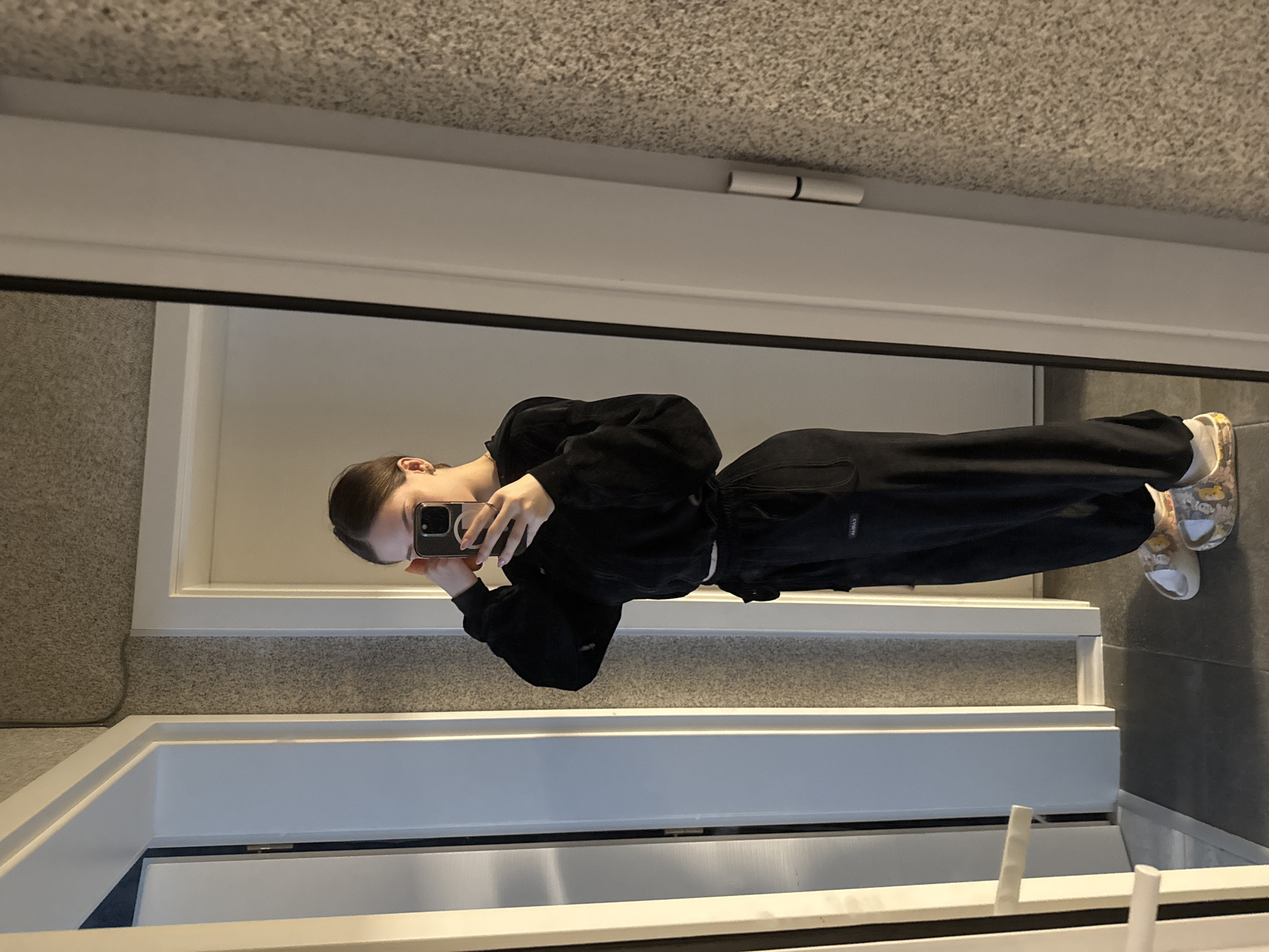 A young woman is taking a selfie in a mirror. She is wearing a black sweatsuit and patterned clogs. The background shows a neutral-toned wall or floor, and the mirror reflects the surroundings. The location is indiscernible as to being Sofia, Bulgaria based on the image information provided.