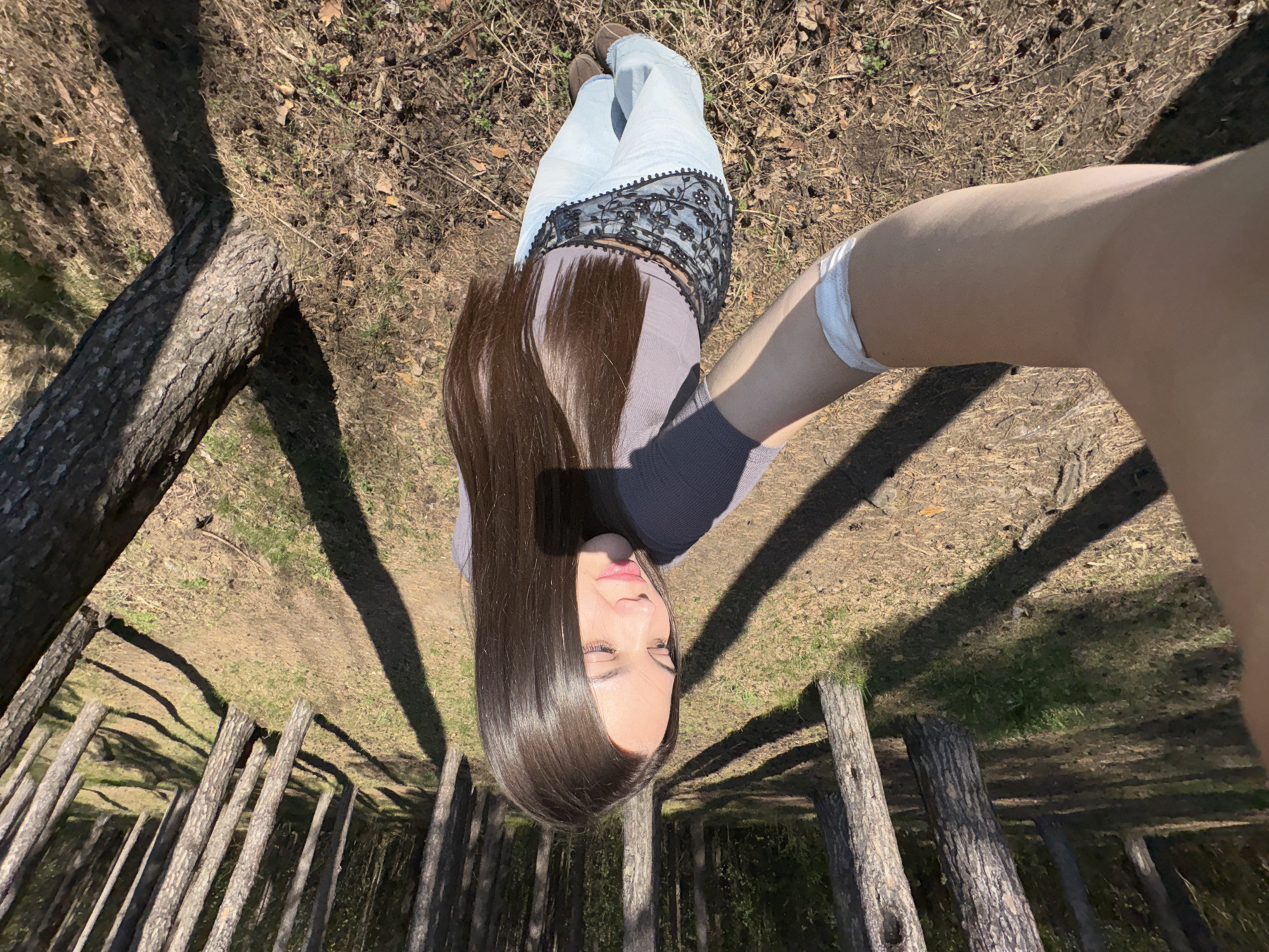 A woman is visible from an overhead perspective, apparently lying on the ground in a wooded area. She is wearing light-colored jeans with a black lace trim, a dark grey top, and brown shoes. Her long brown hair is spread out around her head. Her right arm is extended towards the camera, possibly taking a selfie. In the background, there are several tree trunks and fallen logs, suggesting a forest or park setting. The ground is covered with leaves, soil, and short vegetation. The location is not specified, nor is there any information to identify any city, but the environment appears to be outdoors in a forested or wooded location.