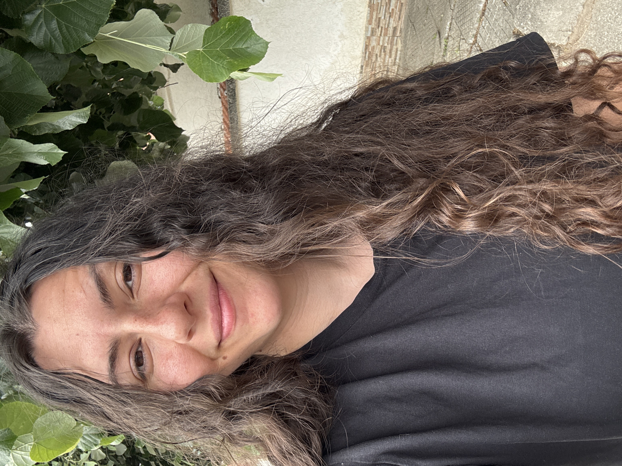 A woman with long, curly brown hair smiles slightly at the camera. She is wearing a black t-shirt. Lush green foliage is visible in the upper left and lower left corners of the image. The background includes a textured wall. The location is Unknown, Unknown.
