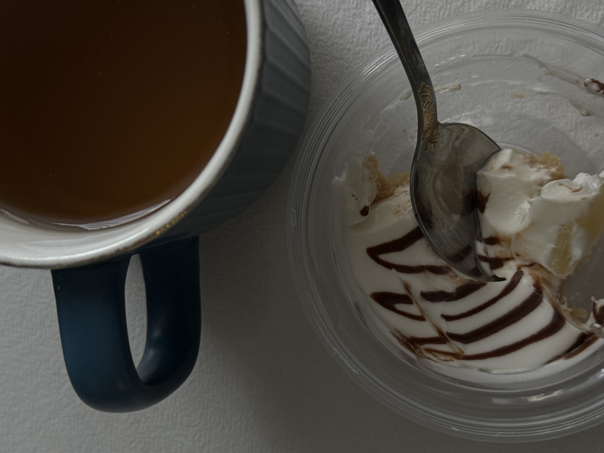 A top-down shot reveals a blue mug filled with a brown liquid, possibly coffee or tea, positioned next to a partially consumed dessert in a clear plastic container. A silver spoon rests within the dessert, which appears to consist of white cream with brown swirls, possibly chocolate sauce, and a layer of cake. The background is a plain, light-colored surface. The location is unknown.