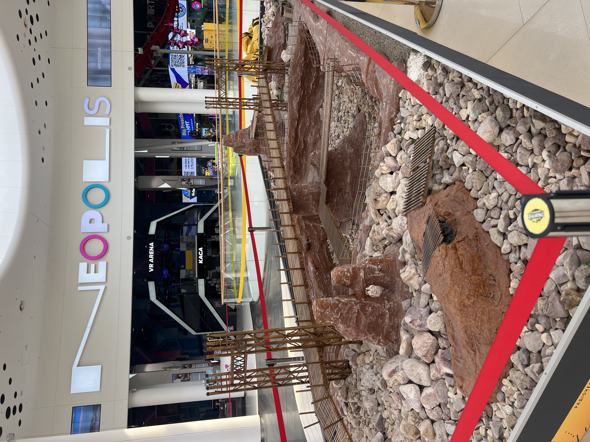 The image depicts a miniature diorama of a rugged landscape with bridges and rock formations, possibly representing a historical or fantasy setting. The diorama is set up in a public indoor space, likely a shopping mall, as indicated by the large signage in the background that reads "NEOPOLIS" and "INTERTOPUA," suggesting it is located in the NEOPOLIS shopping center in Kyiv, Ukraine. The diorama is cordoned off with red ribbon barriers. In the background, various shops and advertisements are visible, including a "VR ARENA" and "KACA" (cashier in Ukrainian). There are no people directly interacting with the diorama. The overall scene suggests a display or exhibit within a commercial environment.