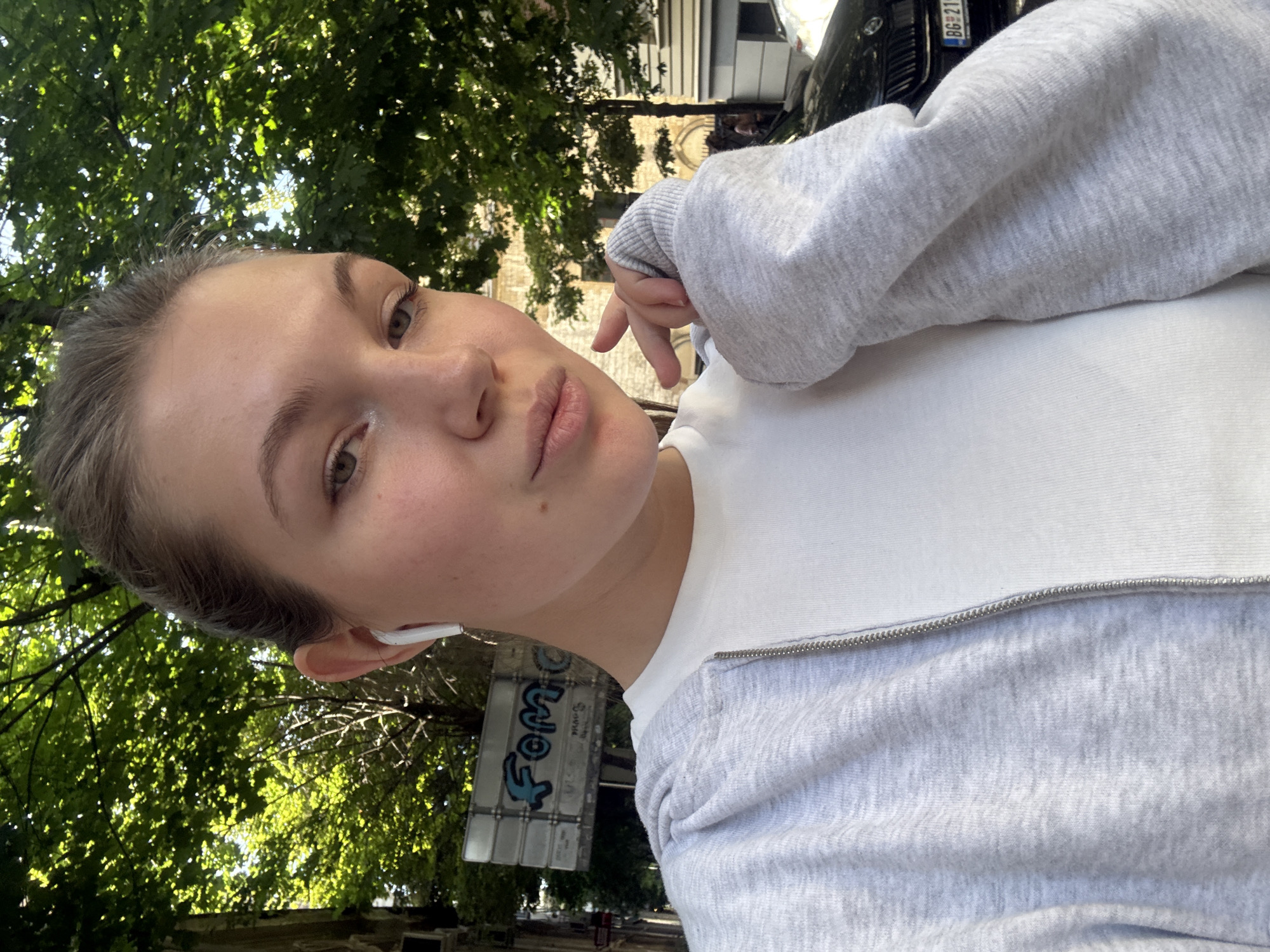 A young woman with short brown hair is taking a selfie outdoors in what appears to be a park or tree-lined street. She is wearing a white shirt under a light gray zippered sweatshirt. She has an earpiece in her left ear. A tree with dense green foliage is behind and to the left of her. In the background, there is a building with a light-colored facade. A black vehicle, possibly a car, with the license plate "BG 21" is visible in the upper right. A street sign with "Fomco" written on it is visible to the lower left. The scene is likely in Belgrade, Serbia.