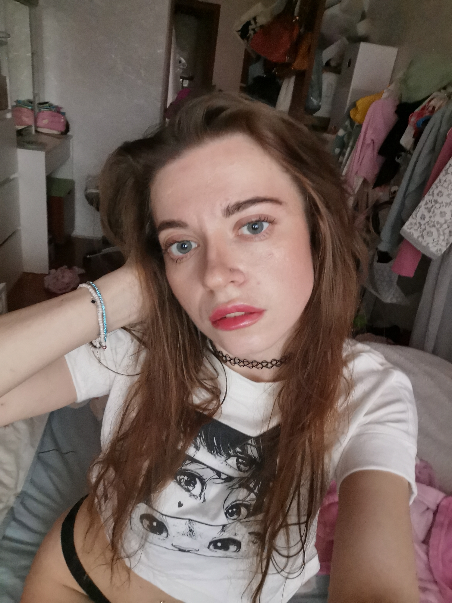 A selfie depicts a young woman indoors. She has fair skin, blue eyes, and auburn hair. The woman wears a white T-shirt with a black anime-style graphic, a black choker necklace, and a beaded bracelet on her left wrist. She is wearing lipstick. The background shows a cluttered room with clothing racks, a dresser, and general disarray. The location is not provided in the city of Unknown, Unknown.