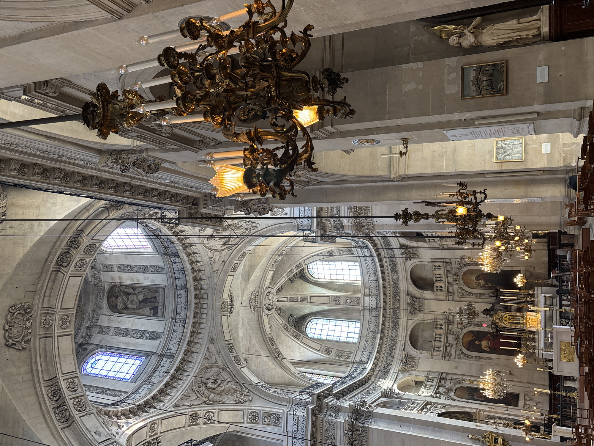 The image depicts the interior of the Saint-Louis-des-Invalides Cathedral in Paris, France. It is a wide-angle shot looking upwards and towards the altar. Dominating the foreground are two large, ornate, gilded chandeliers with numerous candle-like lights. The architecture of the cathedral is grand and neoclassical, with a prominent dome visible in the background, featuring arched windows and decorative moldings. Several other chandeliers and sconces are visible throughout the space. The walls are adorned with paintings and architectural details, including arches and columns. In the distance, the altar area is visible, with religious paintings and an organ. Rows of wooden chairs are arranged in the nave. The lighting is a mix of natural light coming through the windows and the artificial light from the chandeliers, creating a warm and solemn atmosphere. There are no people prominently featured in the image.