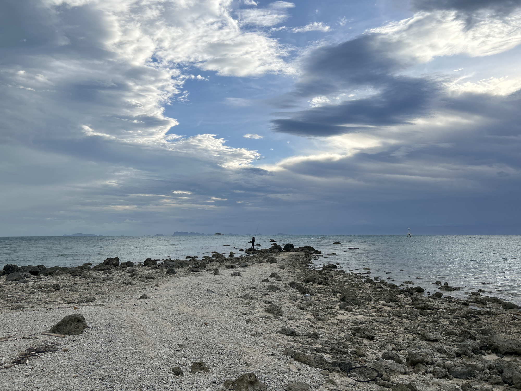 A wide-angle shot captures a rugged, rocky shore leading to the sea under a dramatic, cloud-filled sky. The foreground is dominated by a rough path composed of small white shells and larger grey rocks. To the left, the sea is a calm, light turquoise, meeting a rocky coastline that extends into the distance. In the middle ground, a lone figure, silhouetted against the water, stands on the rocky path, holding a fishing rod. Further out in the sea, a white buoy with a light is visible. The background features a hazy horizon with distant islands. The sky is filled with a mix of dark grey and bright white clouds, with patches of blue sky visible through them, suggesting an overcast or stormy weather pattern.