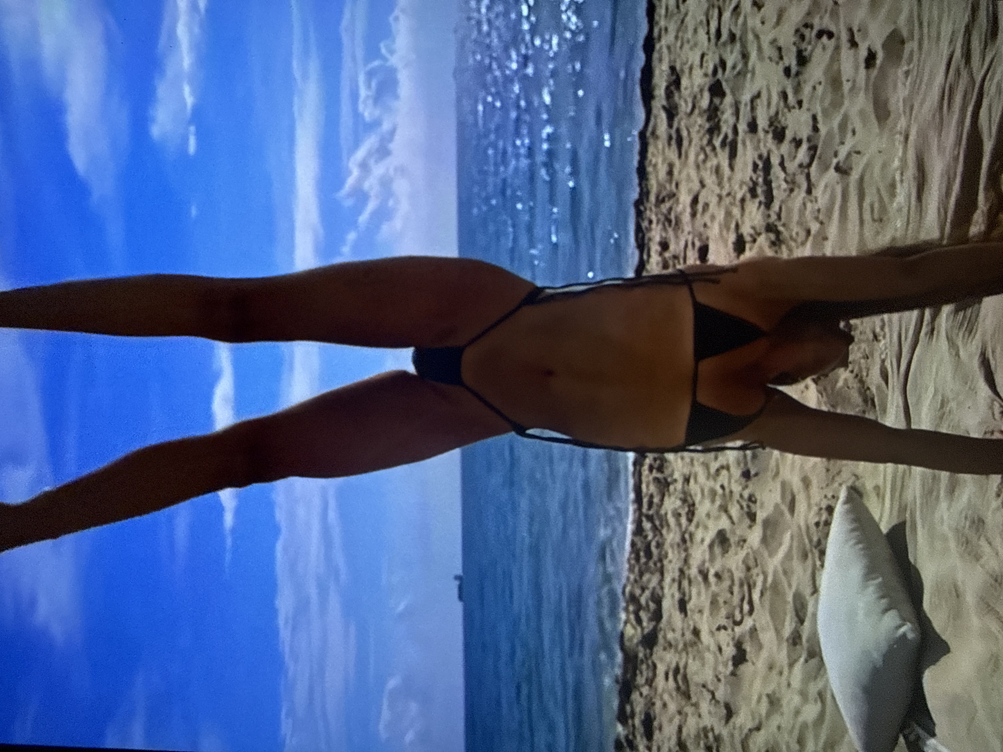 This image depicts a female individual performing a handstand on a sandy beach in Varna, Bulgaria. The person is inverted, supported by their arms with hands placed on the sand, while their legs are extended upwards in a 'V' shape. They are wearing a dark, two-piece string bikini. The background features a vibrant blue sky with scattered white clouds, meeting a blue ocean horizon. A large cargo ship is visible on the left side of the distant ocean. On the far right, faint land is discernible along the horizon. Sunlight reflects brightly off the ocean surface on the right side of the frame. In the lower-left foreground, a white rectangular pillow rests on the sand.