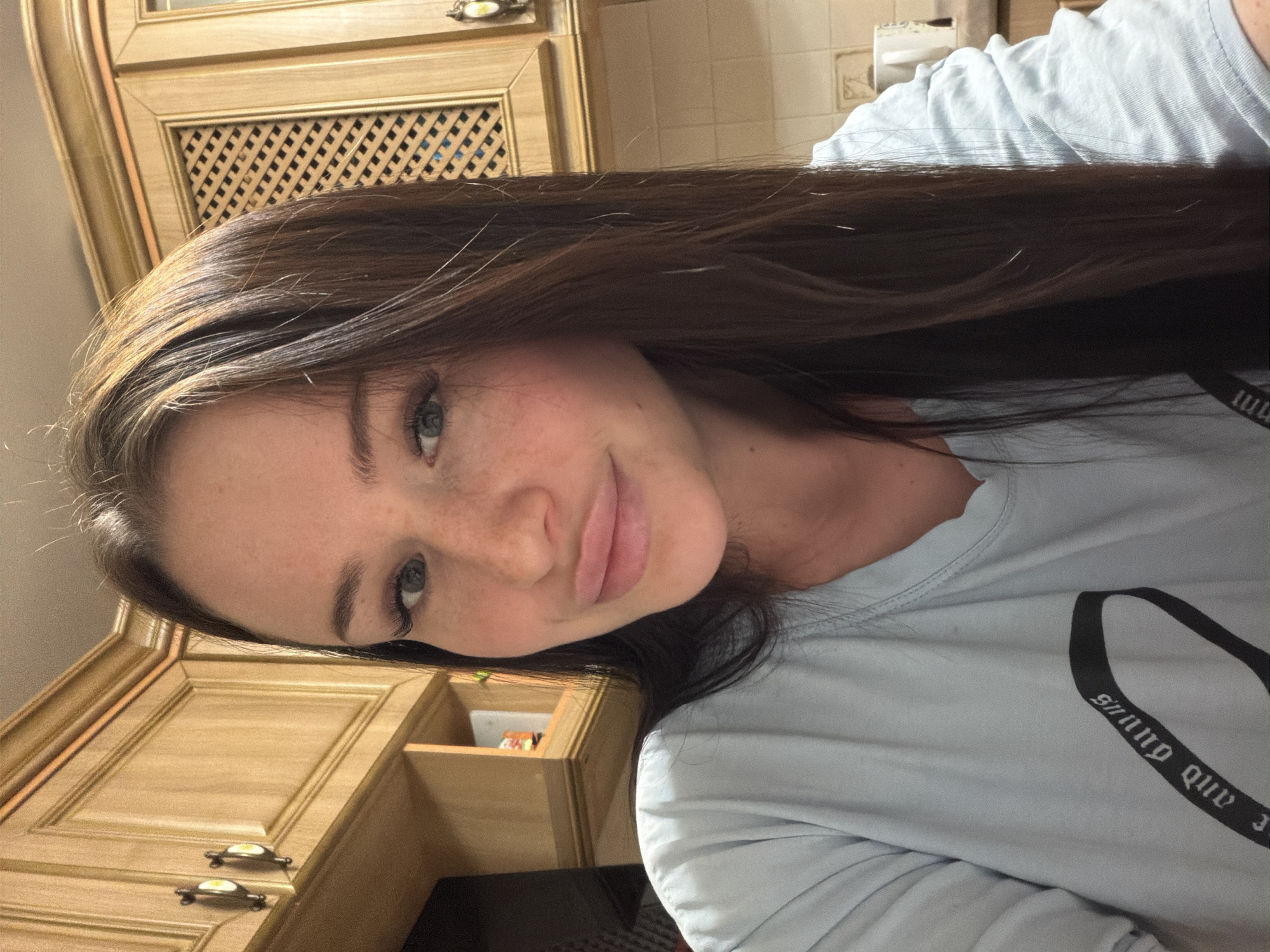 A young woman with long, dark brown hair, fair skin, and light blue-green eyes is the central subject of the image. She has visible freckles across her nose and cheeks and long, dark eyelashes. Her head is tilted, and she is looking directly at the camera with a soft, closed-mouth smile, conveying a calm and pleasant emotion. She is wearing a light blue or light grey long-sleeved t-shirt. The image is rotated, giving the impression that she is lying down.
The setting appears to be indoors, likely a kitchen or dining area. In the background, there are light-colored wooden kitchen cabinets with ornate metal handles. One section of the cabinet features a decorative lattice-patterned insert. Further in the background, light-colored wall tiles and what appears to be a tissue dispenser or similar object are visible on the wall. An open cabinet or shelf, containing some indistinct colorful items, can be seen near her face.
The activity depicted is a close-up portrait or selfie. The lighting suggests a well-lit indoor environment, possibly daytime or bright artificial light.
Visible text on her t-shirt, in a Gothic-style font, reads "gniwD qnd". Due to the likely selfie nature of the photograph and the image's rotation, the text appears inverted and mirrored.