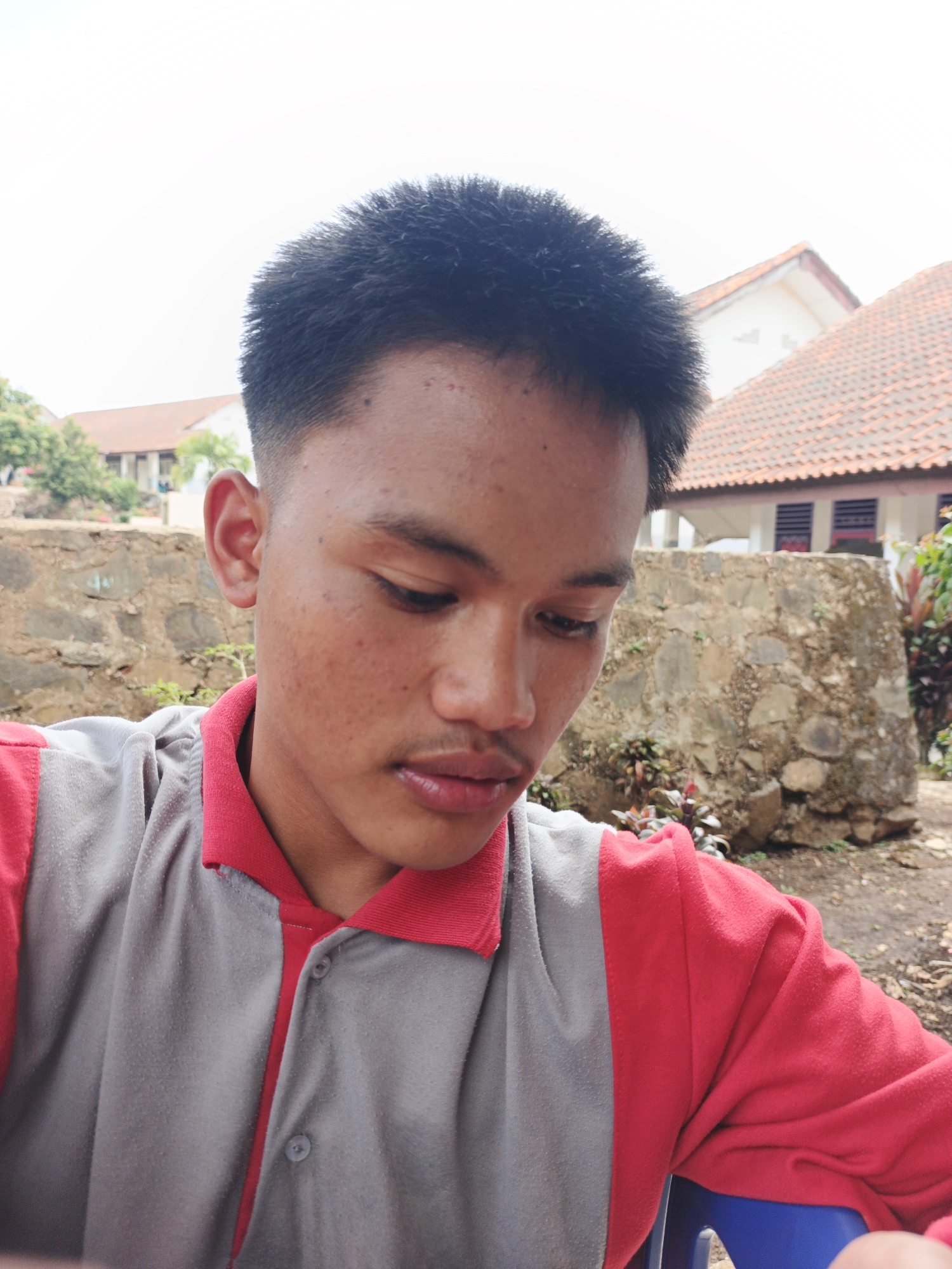 The image shows a close-up, slightly downward-facing portrait of a young male with short, dark hair, facing to the right. He has a fair complexion with visible pores and some blemishes on his face. His eyes are looking down, and his mouth is slightly open. He is wearing a two-toned shirt, with a gray front and red sleeves and collar. The background is out of focus and appears to be an outdoor setting, possibly a village or rural area, with a stone wall, some greenery, and buildings with red tiled roofs. Given the context of Tanjung Setia, Indonesia, this scene could depict a residential area or a school campus within the town.