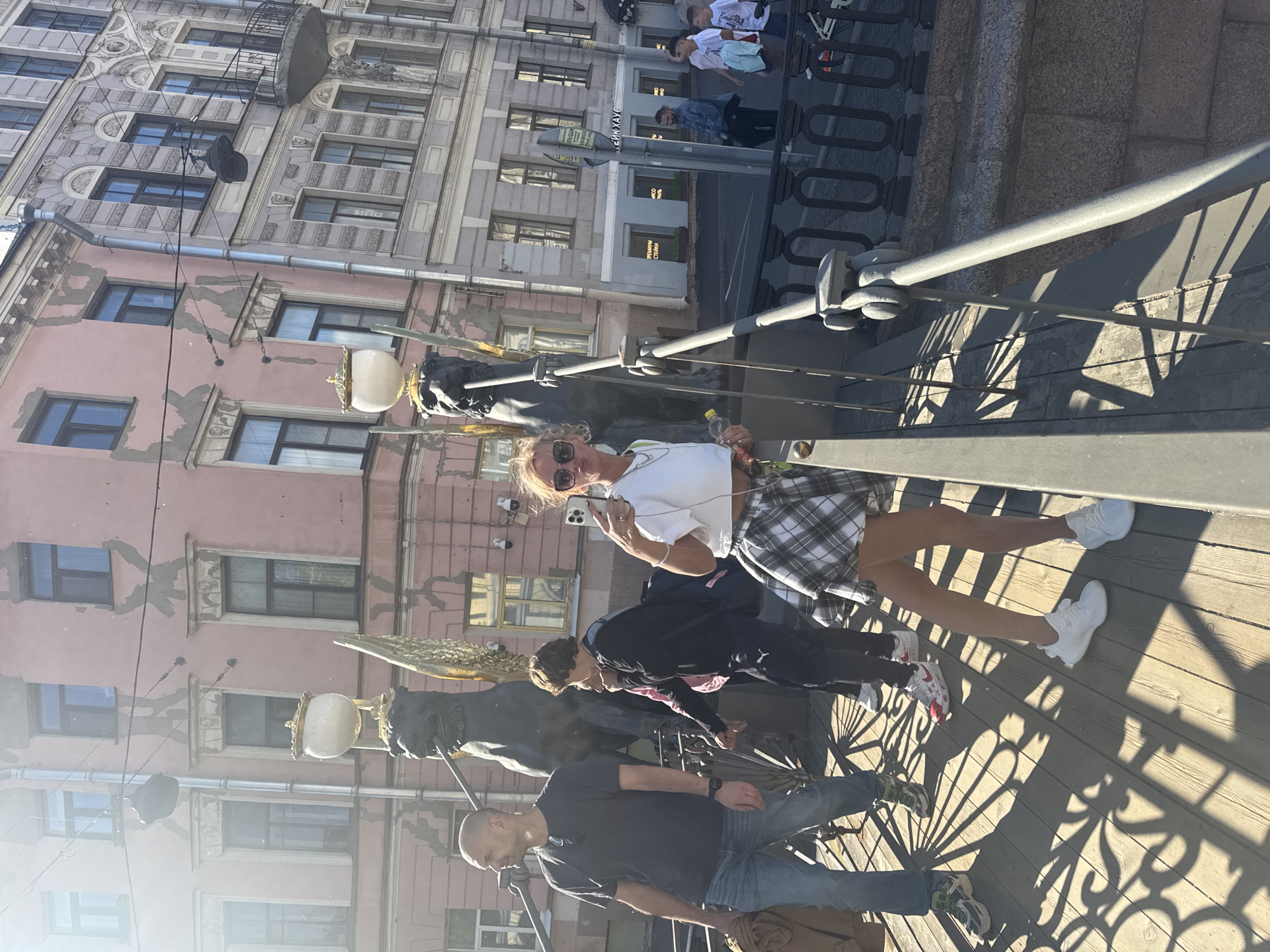 A group of people are on a bridge with a view of buildings in Saint Petersburg, Russia. The bridge has a wooden walkway and ornate metal railings. In the background, two buildings are visible: one is pink with white trim and numerous windows, and the other is grey with similar architectural features. Two griffin statues, often referred to as the "Griffins of Bank Bridge," are situated on the bridge, each holding a lamp.
In the foreground, a woman with blonde hair, wearing sunglasses, a white cropped top, and a plaid skirt, is taking a selfie with her phone. She is holding a plastic bottle in her left hand. To her left, a man with a shaved head, wearing a dark t-shirt and blue jeans, is looking to the left. Behind him, a younger person in a black hoodie and dark pants is also present. Further in the background, on the sidewalk across the street, several individuals are walking. The lighting suggests it is daytime, with strong shadows cast by the bridge's structure and the griffin statues.