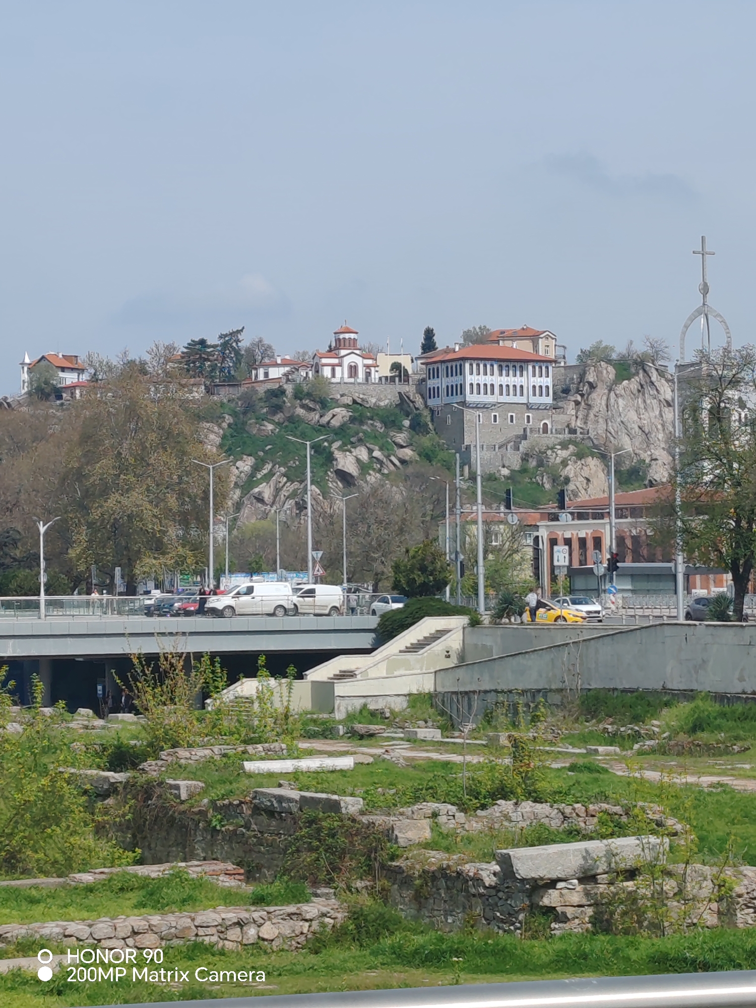 The image captures a daytime urban scene in Plovdiv, Bulgaria. The foreground features partially excavated stone ruins interspersed with patches of grass and low vegetation. A modern concrete structure, possibly a pathway or a section of a building, is situated behind the ruins, with stairs visible.
A bridge spans the middle ground of the image, carrying vehicular traffic, including cars and vans. On the right, there is a yellow taxi.
In the background, a hill dominates the skyline. Structures, including a church with a red-tiled roof and a building with a white facade and red roof, are perched atop the hill, interspersed with rocky outcrops and vegetation. A metal cross stands prominently on the hill. The sky is overcast with a light blue hue.