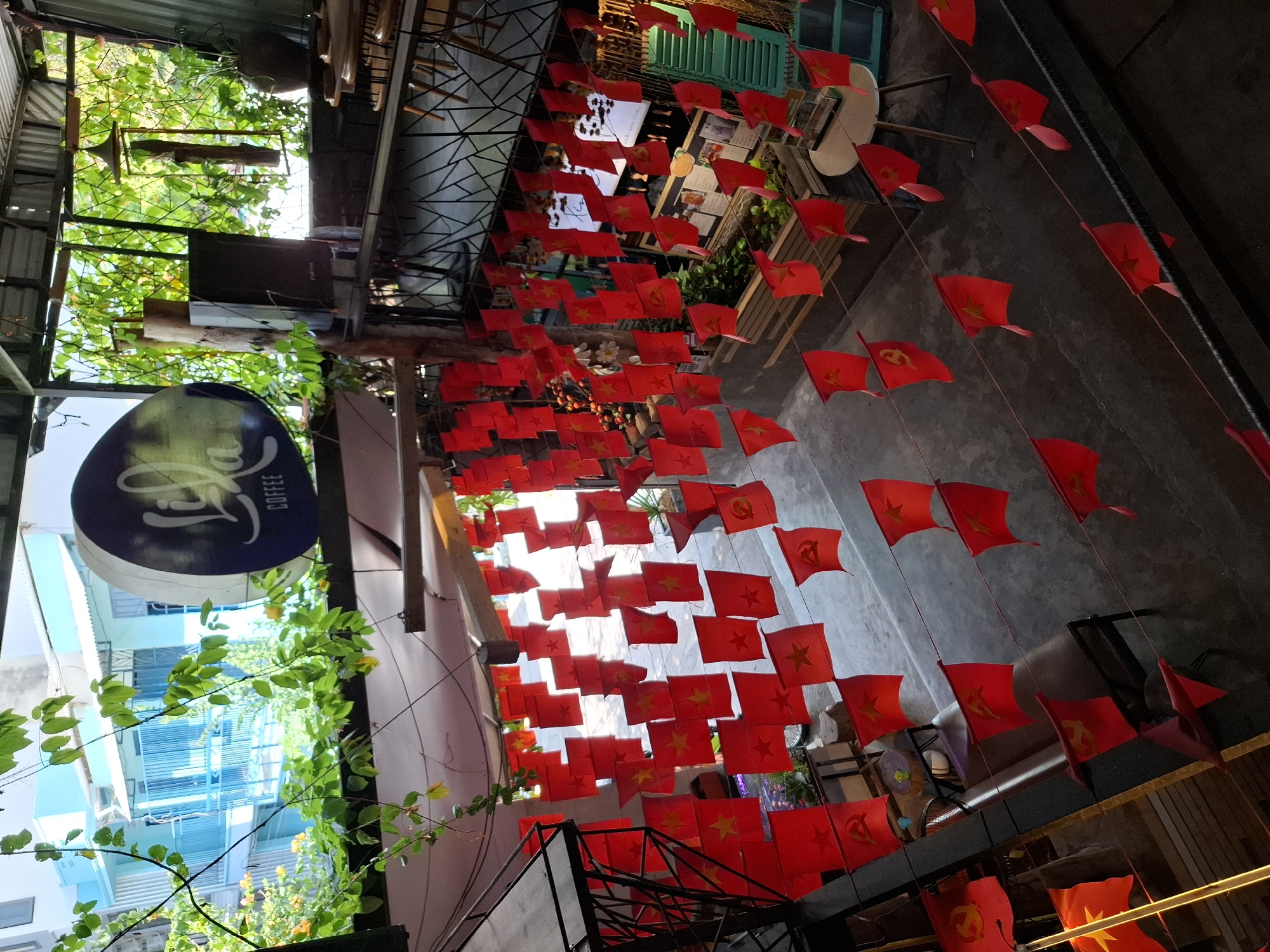 The image depicts the interior of a cafe in Nha Trang, Vietnam, characterized by an abundance of small red flags with yellow stars and hammers and sickles, strung across the space. The flags are arranged in rows and appear to be a form of decoration. The cafe has an open-air feel, with greenery visible outside and an overhead structure made of beams and roofing.
In the foreground, a metal railing and a staircase are visible. The main subject is the extensive display of flags, which hang from wires strung at various heights. The flags are predominantly red, with some featuring a yellow star, while others display a hammer and sickle symbol, indicative of communist or socialist political affiliation. These flags suggest a nationalistic or political theme for the establishment.
Further into the cafe, tables and chairs are set up, some with patrons, though their faces are not clearly visible. There are also potted plants and decorative elements, including a prominent circular sign with text that is partially obscured by foliage but appears to be a logo for "Lila Coffee."
The environment is somewhat rustic, with exposed structural elements and a concrete floor. The lighting suggests it is daytime, with sunlight filtering in from outside. The overall impression is of a casual, perhaps themed, cafe space decorated in a manner that reflects Vietnamese national symbols.