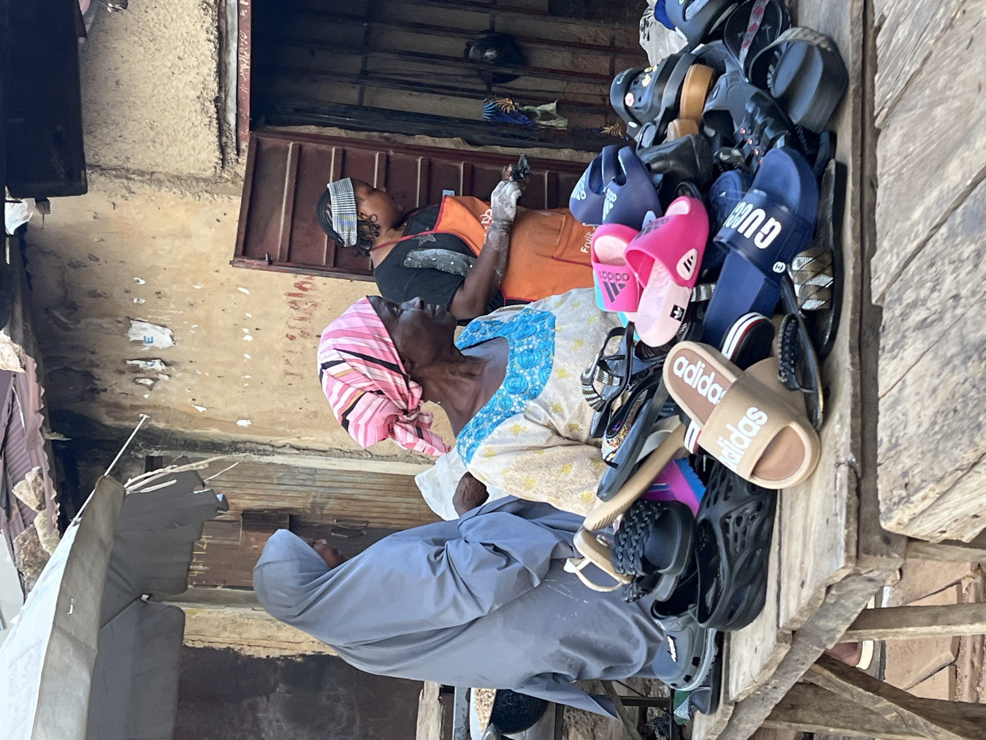 The image depicts a street scene in Jos, Nigeria, where a vendor is selling footwear from a wooden stall. Three women are present. The woman in the foreground, wearing a pink and white headscarf and a light-colored patterned top with a blue neckline, is seated and appears to be interacting with the footwear. To her left, another woman in a grey hijab is standing and looking away from the camera, partially obscured by the stall's umbrella. To the right, a third woman, wearing a black top and an orange apron, is standing and looking down, seemingly engaged in an activity with her hands, possibly attending to the merchandise. The stall is piled high with various types of sandals and flip-flops, including brands like Adidas and Gucci, in a range of colors. The background shows a building with weathered walls and a corrugated iron roof, typical of the urban environment in Jos. The overall impression is that of a busy marketplace with a focus on informal retail.