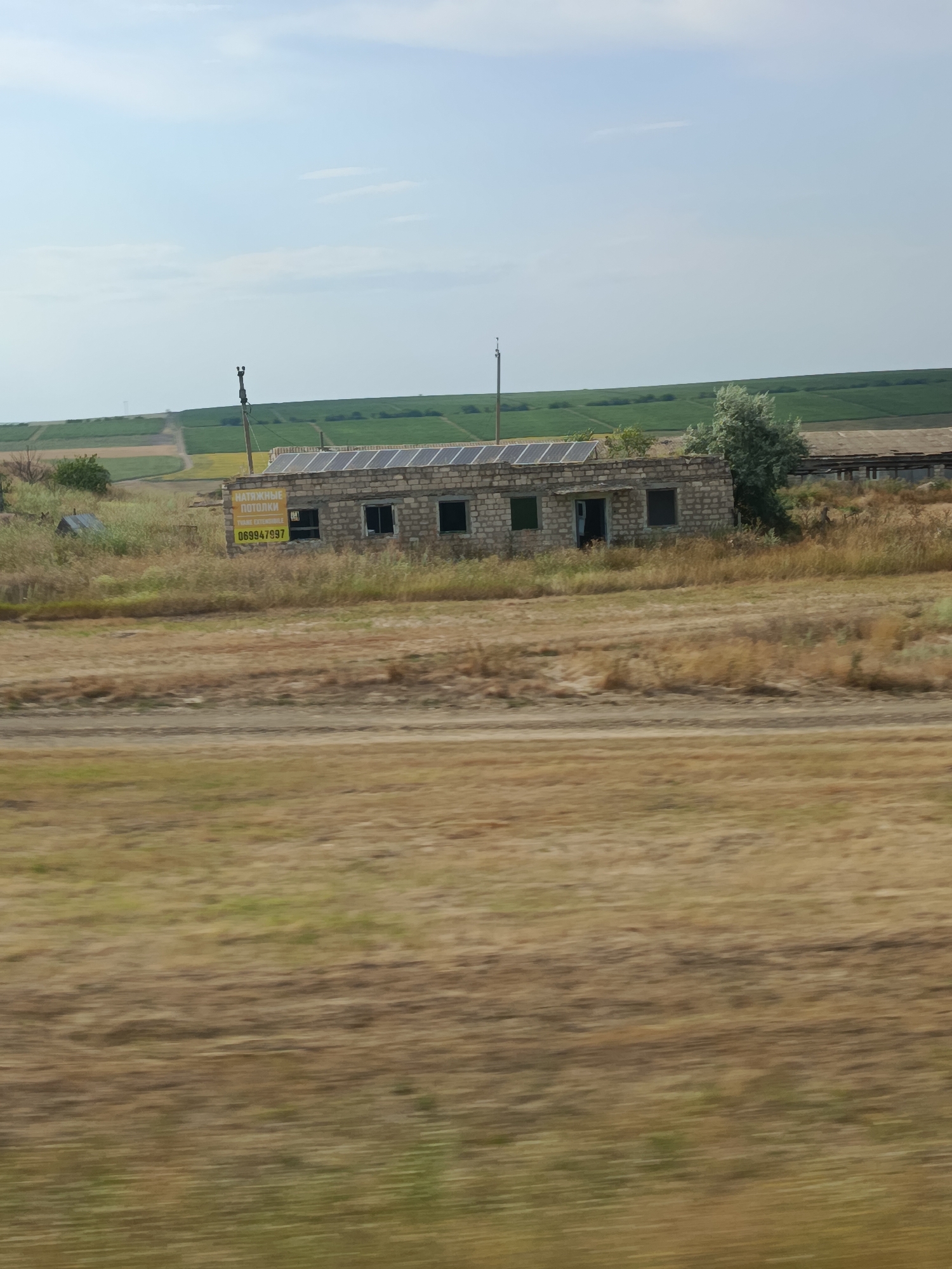 A low-angle, slightly blurred shot captures an abandoned brick building with solar panels on its roof, situated in a rural landscape near Comrat, Moldova. The building has a partially open doorway and several windows, some of which appear to be broken or boarded up. A yellow sign with Cyrillic text and a phone number, "069947997", is affixed to the front of the building. The surrounding environment is characterized by dry, golden grass and fields in the background, suggesting a dry season. In the distance, green agricultural fields and more structures can be seen. A utility pole stands to the left of the building, and a tree with silvery-green foliage is located behind it. The sky is a pale blue with wispy clouds. The overall scene evokes a sense of abandonment and disuse in a agricultural setting.
