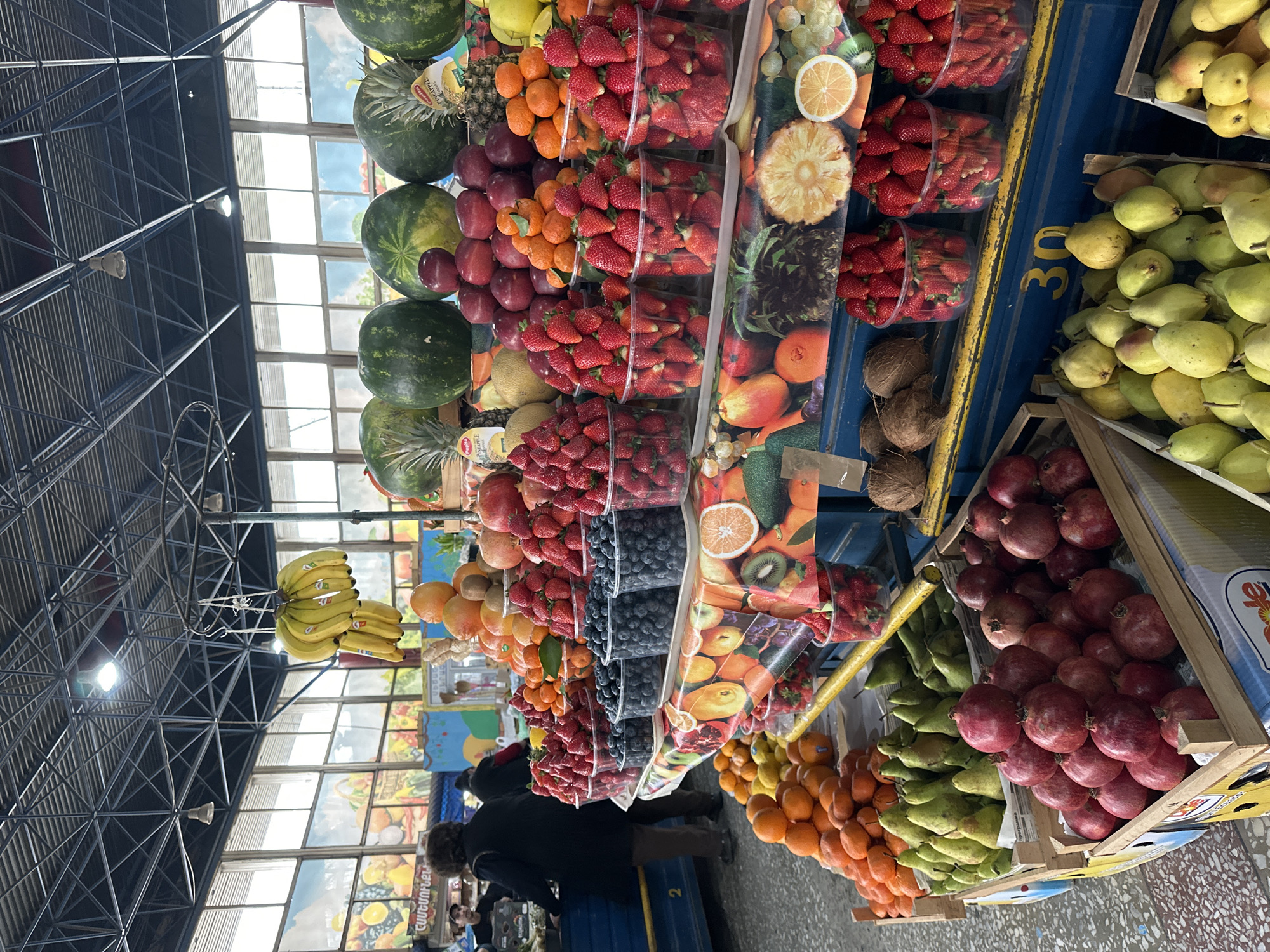 This image depicts a vibrant fruit market scene. The market is located in Yerevan, Armenia, identified by the Armenian text and signage visible. The produce stall is overflowing with a colorful array of fruits: watermelons, pineapples, oranges, strawberries in plastic containers, apples, blueberries, bananas, pomegranates, and pears. Several people are seen browsing the produce, likely customers. The market appears to be indoors, characterized by a complex metal framework supporting the roof and windows along the upper part of the wall.