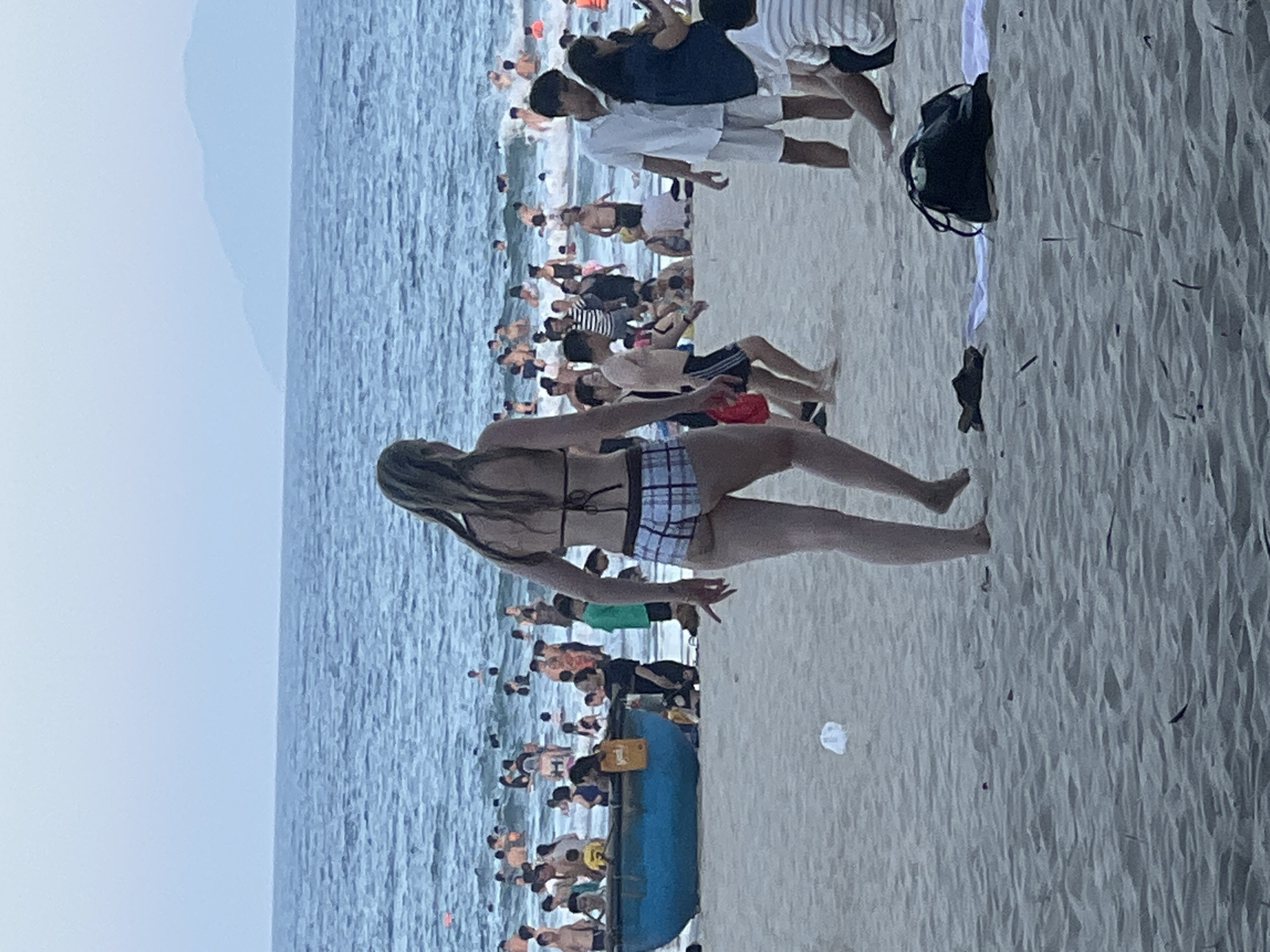 The image depicts a crowded beach scene in Đà Nẵng, Vietnam. The foreground shows sand with a black bag and a towel. In the middle ground, a woman wearing a checkered bikini is walking away from the camera, with her back partially visible. Behind her, numerous people are in the ocean, swimming and playing in the waves. A blue boat is also visible on the beach, with several individuals inside and around it. In the background, the ocean extends to the horizon, with faint mountains visible in the distance under an overcast sky. The overall impression is a lively and active beach environment.