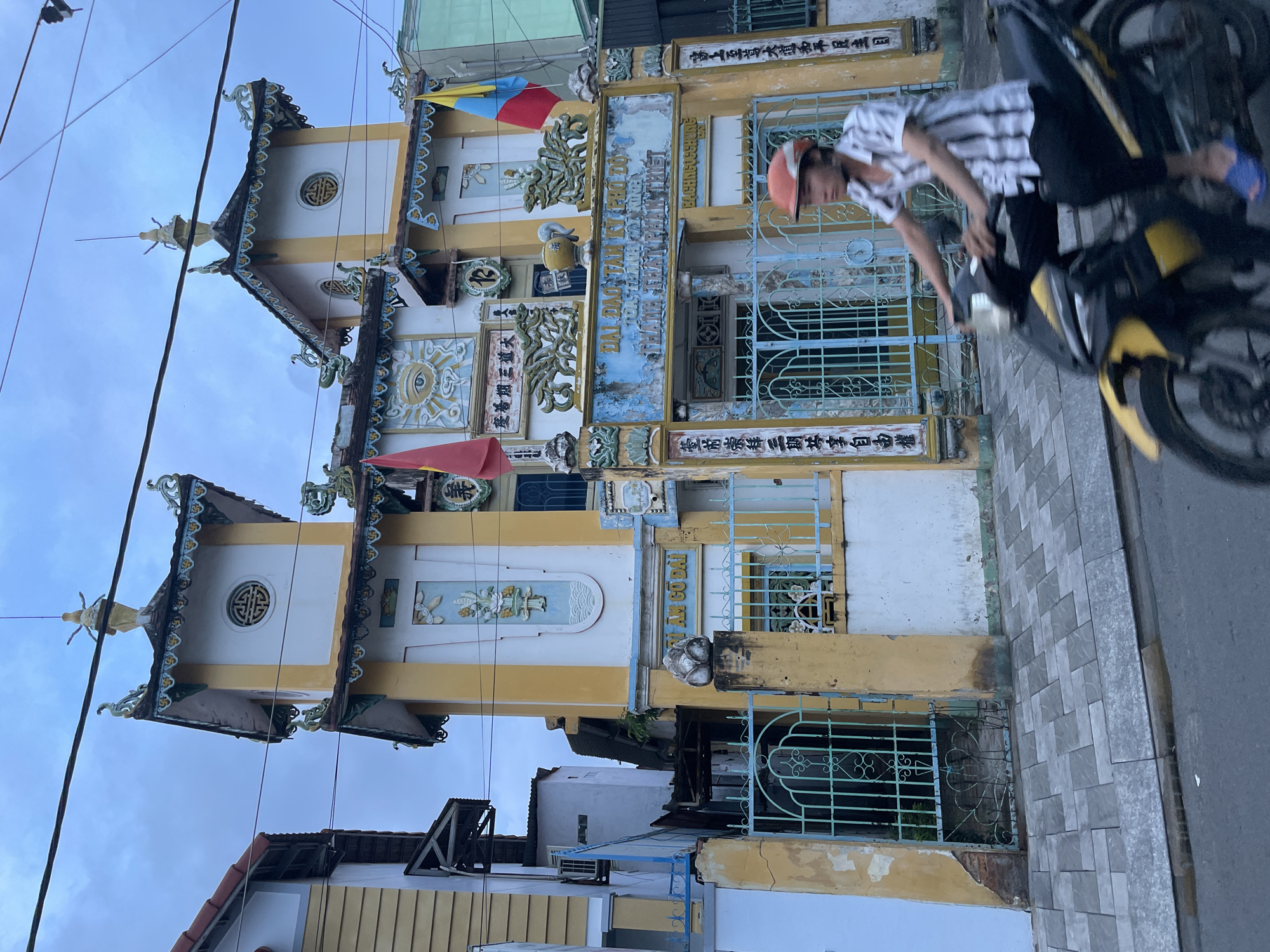 A street scene in Phan Thiết, Vietnam, captures a unique, ornate building under an overcast sky. In the foreground, a person wearing an orange helmet and a striped shirt sits astride a yellow and black motorbike, facing towards the right. The motorbike is positioned on a paved sidewalk next to a road, with the rider's hands on the handlebars.
The dominant feature of the image is the multi-story building behind the person. It is characterized by vibrant yellow and white walls, intricate blue and green decorative carvings, and distinct tiered roofs with upturned eaves, suggesting a temple or pagoda. Two prominent tower-like structures with circular, patterned windows rise from the building. One roof features a small golden statue at its pinnacle. The facade is richly decorated with bas-reliefs, including a large eye symbol radiating light, and various other ornamental motifs. Small red and yellow-blue flags adorn the upper section. The ground floor has blue-green metal gates and window grilles.
Several inscriptions are visible on the building. Text identified includes: "ĐẠI ĐẠO TAM KỲ PHỔ ĐỘ" (The Great Way of the Third Period of Redemption), "BẠCH NGỌC CHUNG", "QUANG MINH CỔ ĐẠI", "TRÂN TRỌNG SÙNG BÁI TAM KỲ CÔNG GIÁI TỰ DO QUYỀN", and "CỨU THẾ TÔN ĐẠI ĐẠO HÒA BÌNH DÂN CHỦ MỤC". These phrases indicate the building is likely a Caodaist temple, given the prominence of "Đại Đạo Tam Kỳ Phổ Độ." Overhead, electrical wires crisscross the frame, adding to the urban setting.