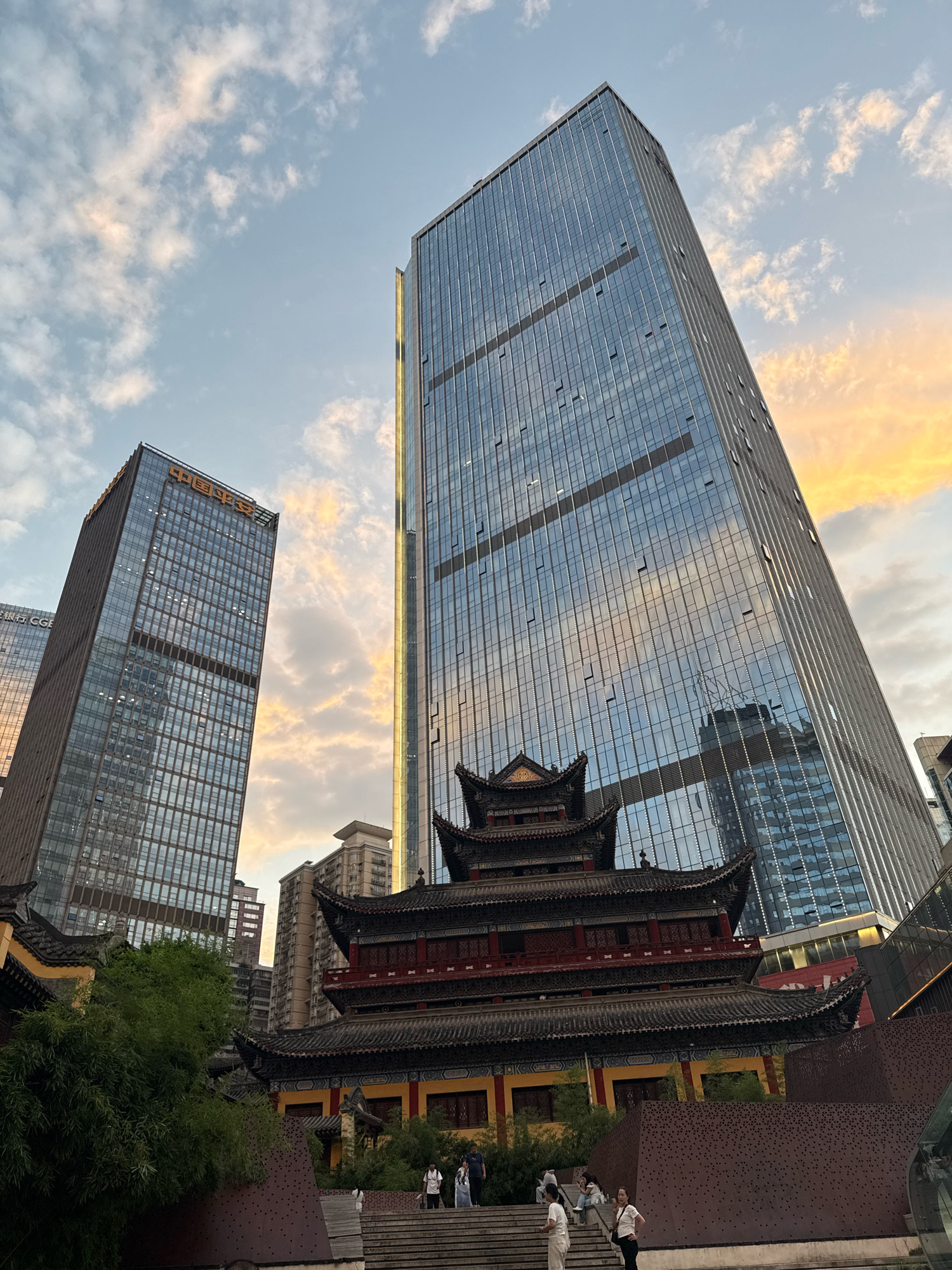In the Yuzhong District of China, a photograph captures a striking juxtaposition of traditional architecture and modern skyscrapers under a sky filled with scattered clouds tinged with the warm hues of sunset. In the foreground, a multi-tiered traditional Chinese temple, adorned with dark tiled roofs and red and yellow detailing, stands prominently. Steps lead up to the temple, and several individuals are seen ascending or standing on them. To the left and right of the temple, towering glass skyscrapers dominate the skyline, their reflective surfaces mirroring the sky and surrounding buildings. One skyscraper on the left features Chinese characters at its apex, likely indicating a company name. The overall scene suggests a dynamic urban environment where historical sites are preserved amidst contemporary development.