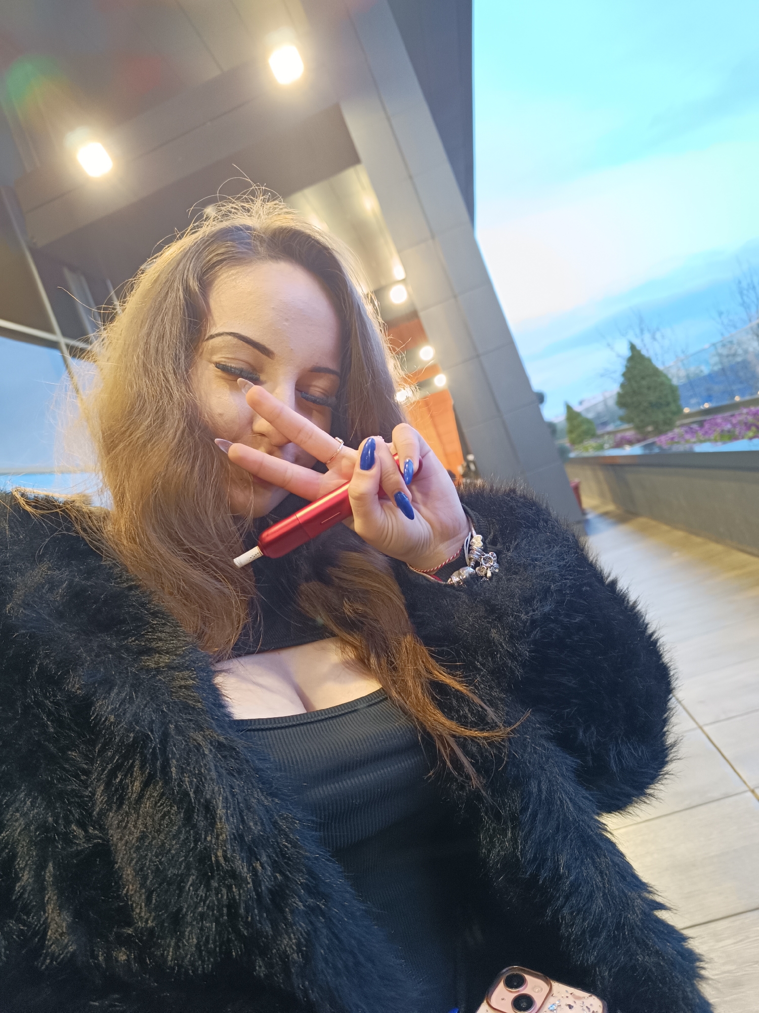 A medium eye-level shot captures a woman outdoors in what appears to be a modern urban setting. The woman, likely in her late 20s or early 30s, is posing with her right hand in a peace sign near her face. She holds a red electronic cigarette in her hand with manicured blue nails. She has long brown hair, fair skin and her eyes are partially closed. She is wearing a black top and a fuzzy black coat.
In the background, the image shows architectural structures with straight lines and modern design. There is a view of a blue sky and some distant greenery including trees and colorful flowerbeds, which appear to be on an upper-level terrace or balcony. Given the presence of specific landmarks such as the modern architecture, the scene may be in or near the city of Plovdiv, Bulgaria. A phone is visible at the bottom right of the image.