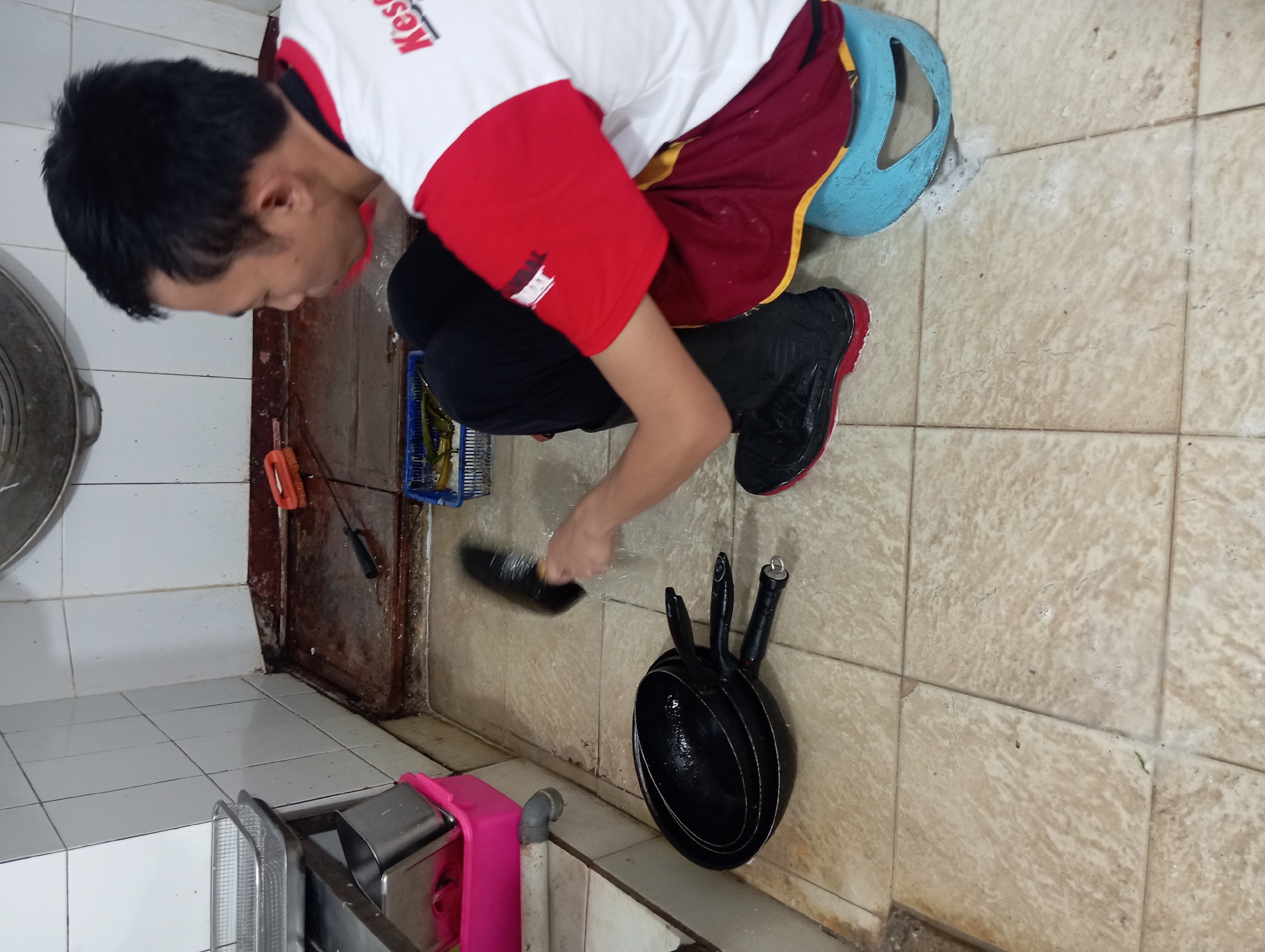 A man is washing three black frying pans in a kitchen. He is wearing a white and red shirt, black pants, and black boots. He is sitting on a blue stool. The kitchen has tiled walls and a tiled floor. There are various cleaning supplies and containers in the background. The scene appears to be in a commercial kitchen.