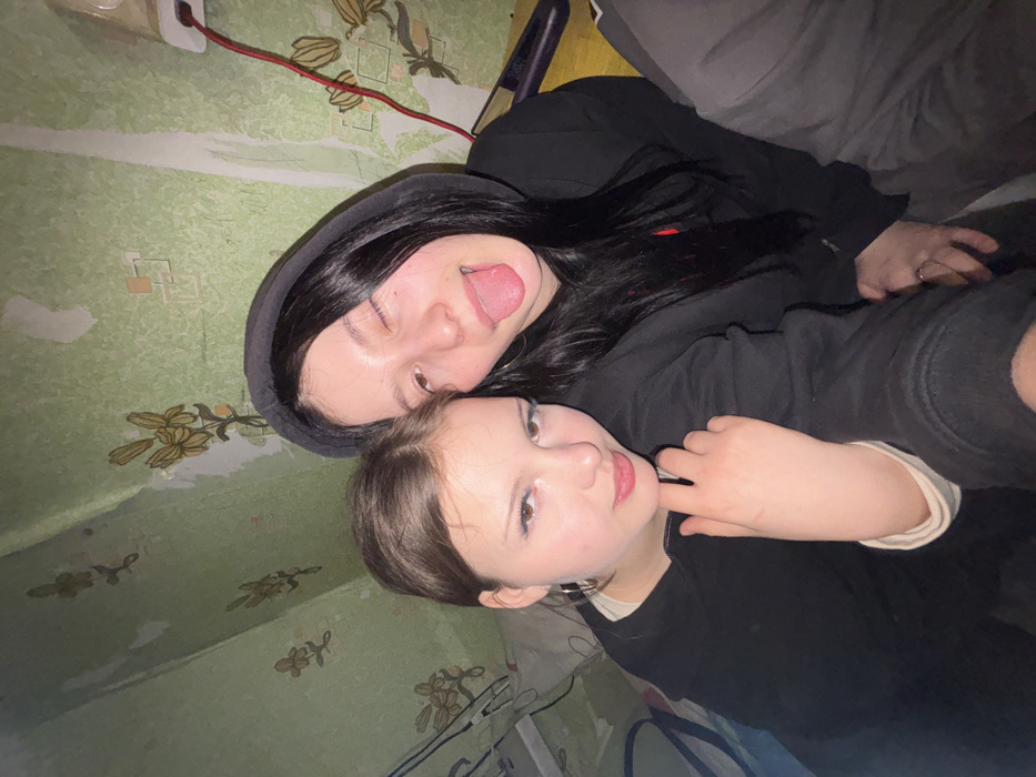 Two young females are depicted in a close-up selfie-style image, taken indoors in the city of Фоки, Russia. The female on the left has light brown hair, wears a dark long-sleeved top over a lighter-colored garment, and has visible hoop earrings and blue eyeliner; her right hand is positioned near her chin, and she displays a subtle smile towards the camera. The female on the right, with long dark hair under a black hooded sweatshirt, is winking with her tongue extended. She is positioned slightly behind and to the right of the first female. A third individual, wearing grey pants, is partially visible on the far right. The background consists of a wall covered in worn light green wallpaper featuring a repeating pattern of stylized yellow flowers with green stems. The wallpaper exhibits several areas of tearing and discoloration. In the upper right corner, a white electrical outlet is visible, with a red cable plugged into it and extending downwards. A dark electronic device is partially visible on a surface below the outlet.