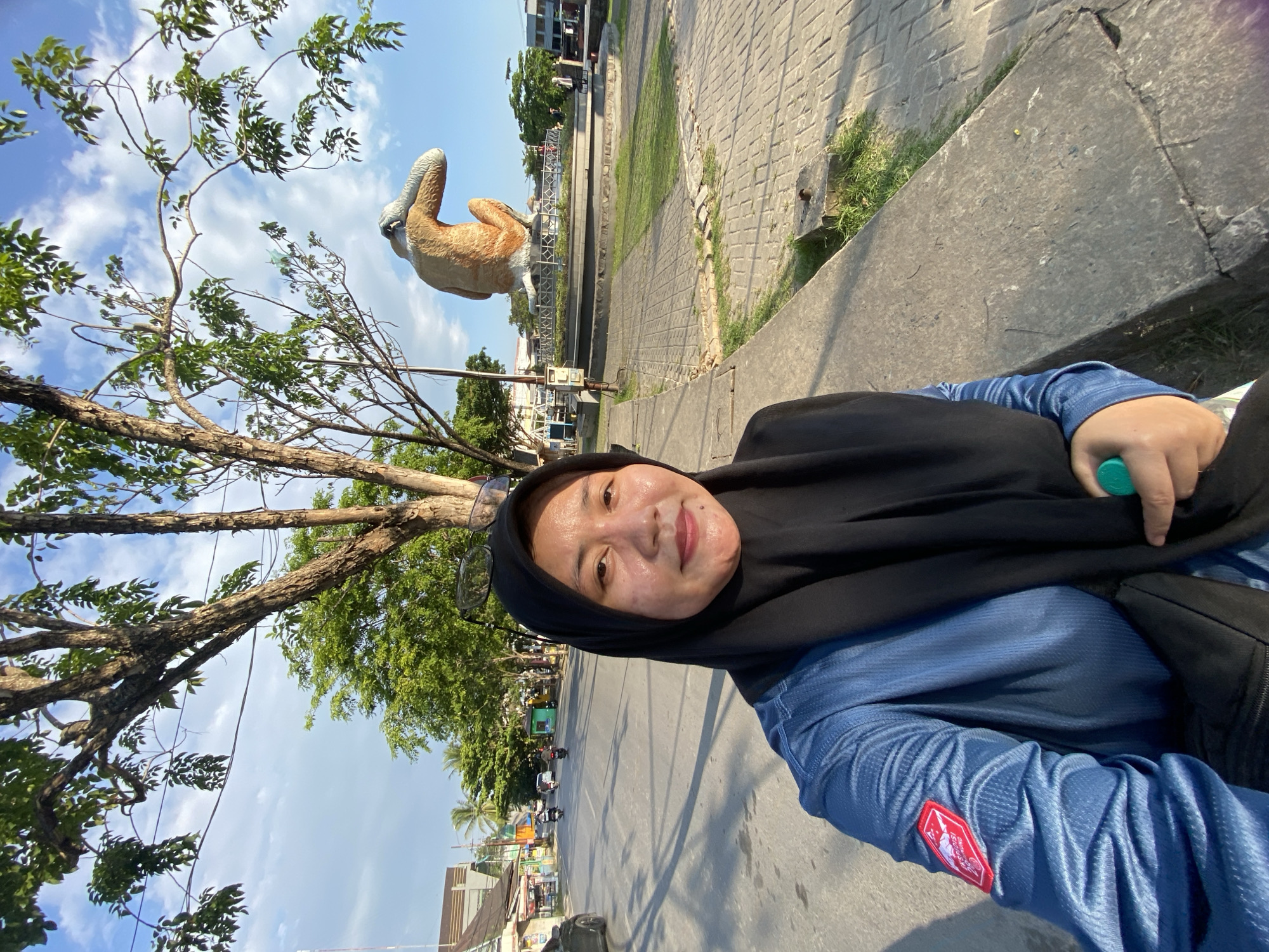 A woman in a black hijab and blue long-sleeved shirt takes a selfie in front of a large statue of a proboscis monkey. The statue is brown and orange and sits on a circular pedestal with decorative fencing and a grassy area. The woman is smiling at the camera and holding a green object in her right hand. In the background, there is a street with traffic, including motorcycles and cars, lined with buildings and trees. The sky is blue with scattered clouds. The scene appears to be in Banjarmasin, Indonesia, a city known for its proboscis monkey population and related tourism. The lighting suggests it is daytime, possibly late afternoon due to the long shadows.