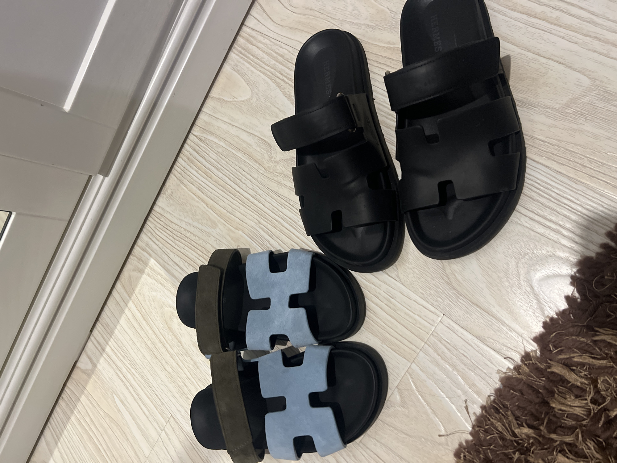 The image shows four pairs of designer sandals laid out on a light-colored wooden floor. Two pairs are on the left, featuring a black base with blue H-shaped straps and a darker strap across the top. The other two pairs are on the right, entirely black, with similar H-shaped straps. A piece of dark brown, shag carpet is visible in the lower left corner of the frame. The background shows a portion of a white, paneled wall, possibly a closet door or room divider. The image does not contain any information that can be used to determine its location in Chișinău, Moldova.