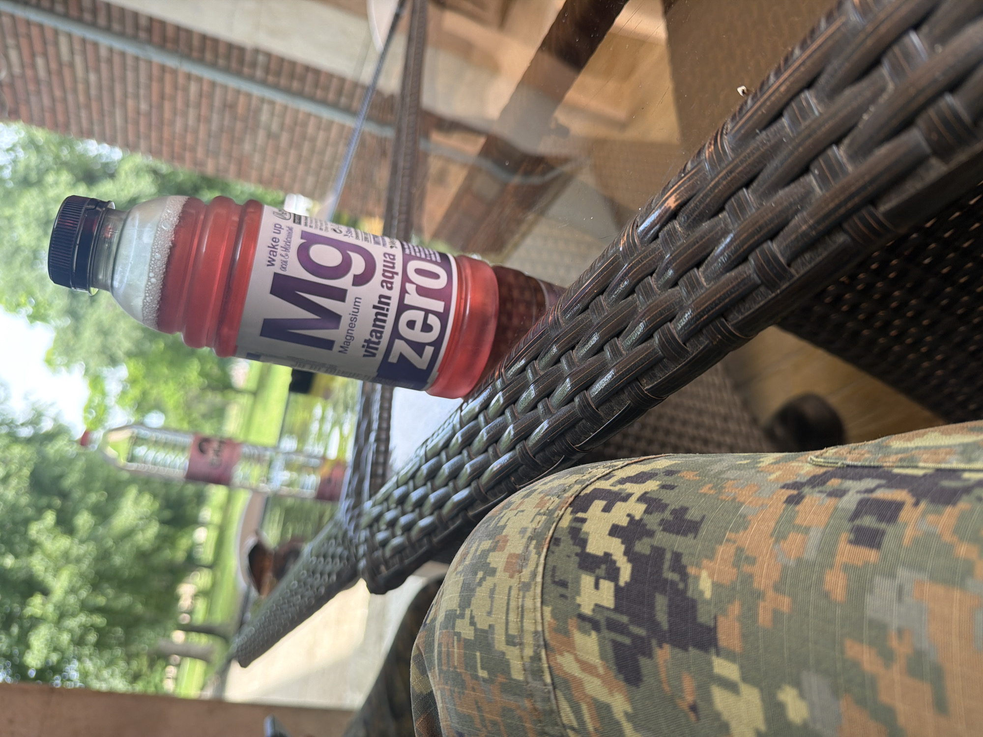 A plastic bottle of Magnesium Vitamin Aqua Zero drink rests on a brown wicker table. Camouflage patterned clothing is visible in the lower right of the image. The background shows trees and a brick building. There is no indication that this image was taken in Dolna Mitropolia, Bulgaria.
