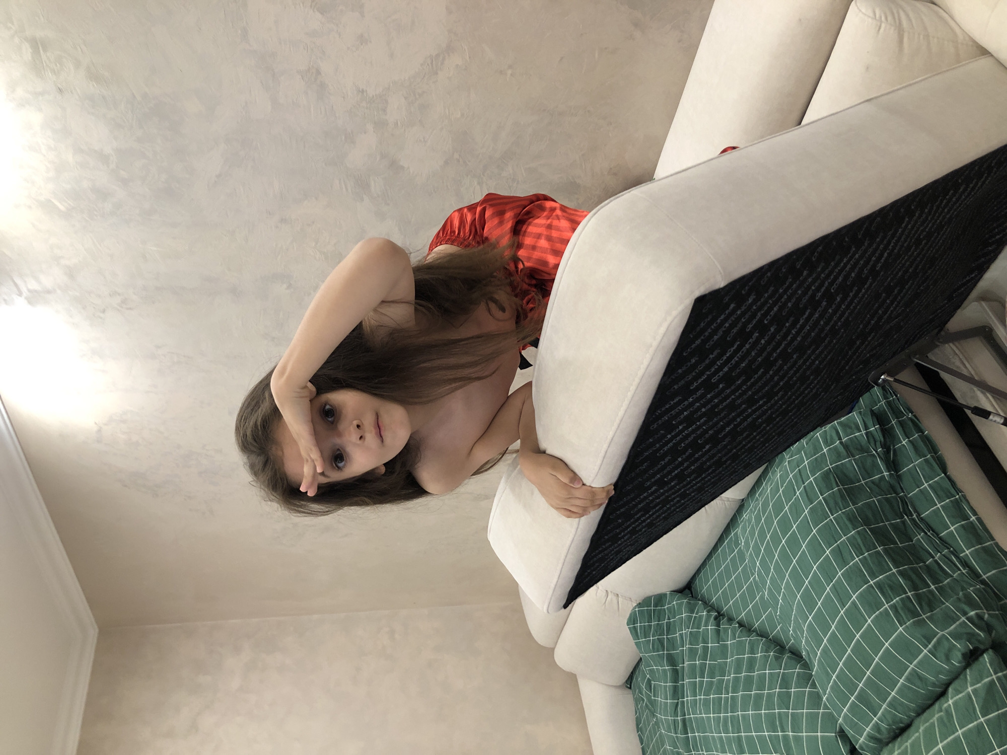 The image displays a young girl, possibly a child, with long brown hair and dark eyes, looking directly at the camera. She is positioned as if peering over or leaning on a large, white sofa. Her left hand is raised to her forehead, shielding her eyes, while her right hand rests on the sofa cushion. She is wearing a red garment with subtle stripes, visible on her back and shoulder.
The background features a light-colored wall with a textured, stucco-like finish, suggesting an indoor setting, likely a home in Saint Petersburg, Russia. The white sofa, possibly upholstered in fabric, occupies a significant portion of the right side of the frame. A green blanket with a distinctive white grid pattern is draped over the sofa.
The lighting is bright and even, indicative of daytime. The girl's expression appears curious and focused, possibly playful. Visible on the black fabric underneath the sofa cushions, the word "COMFORTONOVA" is repeatedly printed in white. The overall impression is a candid, informal moment captured indoors.