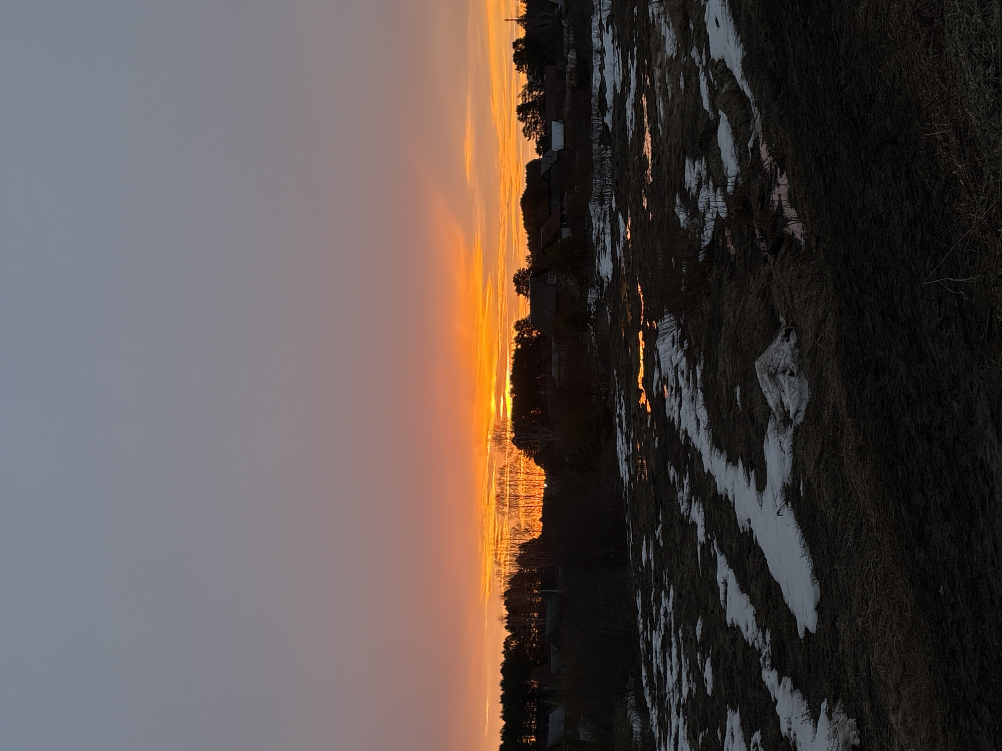 The image shows a sunset in Rusalevka, Russia. The sky transitions from gray in the upper left to a vibrant orange along the horizon. Silhouetted trees and buildings are visible against the orange sky. The ground is covered with patches of snow and dry grass. Pools of water reflect the colors of the sunset. There are no people visible in the image.