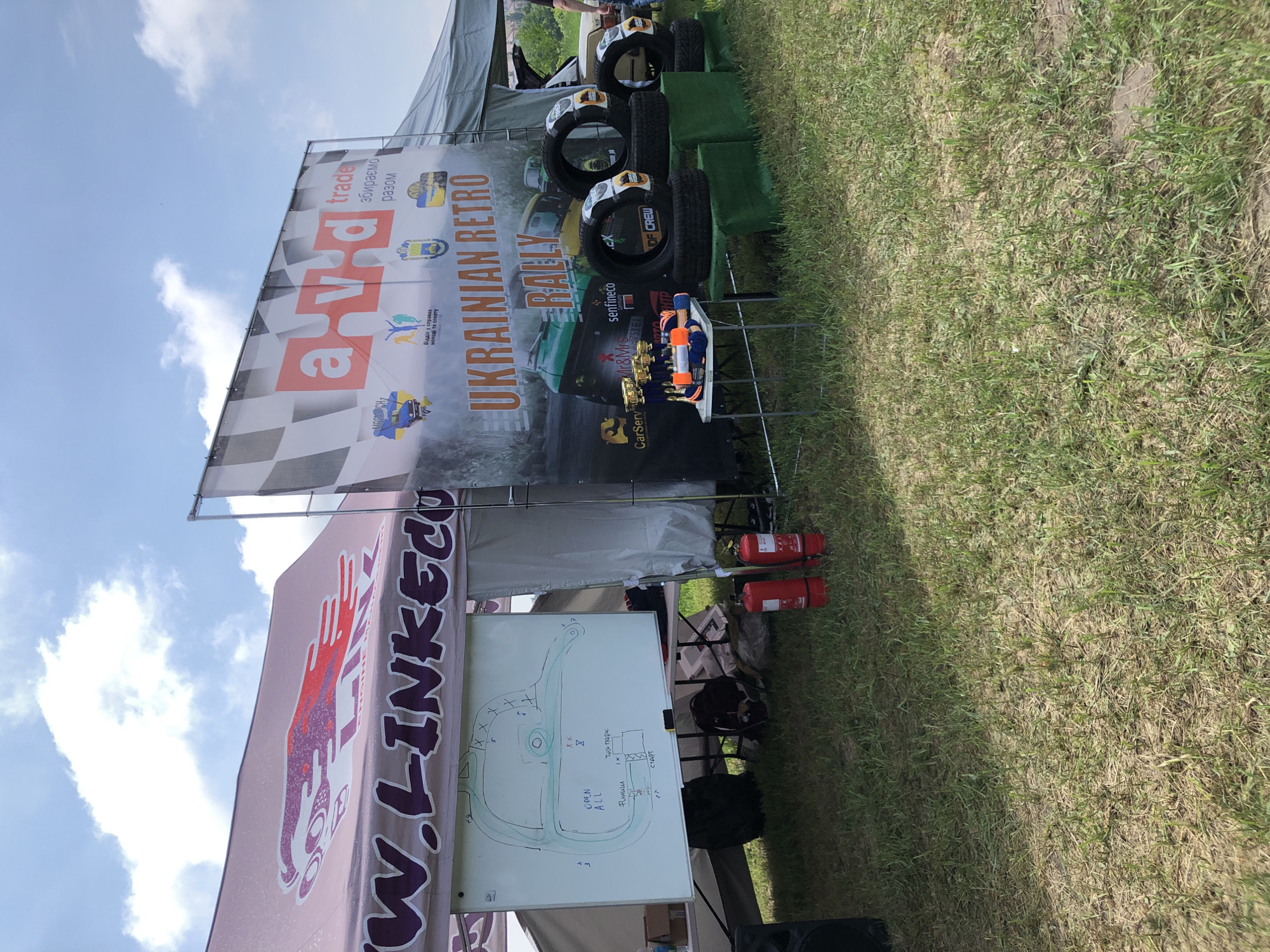 The image depicts an outdoor event taking place on a sunny day in an unknown outdoor location in Unknown, Unknown. The scene features two adjacent canopies and a large promotional banner, set on a patch of sparse, dry grass.
To the left, a pink canopy tent is visible, bearing a large dark purple logo with stylized lettering "W.LINKED.CO" and a red graphic resembling a stylized creature, possibly a jaguar, with the word "ENGINE" partially visible beneath it. Underneath this canopy, a white magnetic board stands on an easel, displaying a hand-drawn diagram in green and black marker. The diagram appears to be a race track or course layout, annotated with text in Ukrainian such as "СТАРТ" (START), "OPEN ALL," and "Тех парк" (Tech park), along with various numerical and symbolic markings. On the ground near the whiteboard, two red fire extinguishers are placed, alongside a black backpack and part of a dark speaker.
To the right of the pink canopy, a large vertical banner is prominently displayed. The banner features a white and black checkered pattern in its upper left corner. A large red "a-v-d" logo is centered, accompanied by the word "trade" and Ukrainian text "збираємо разом" (collect together). The main title, "UKRAINIAN RETRO RALLY," is displayed in large orange letters. Below the title, images of two vintage cars, one green and one yellow, are depicted. Various sponsor logos are arranged at the bottom, including "CarServ," "Mr&Mrs," "senfineco Germany," and "DF CREW." Above the main title, smaller graphical emblems include one featuring a car and the Ukrainian flag.
In front of the banner, a small silver folding table holds multiple gold-colored trophies and several orange, cylindrical objects, possibly small cones or additional awards. Adjacent to the table, several vehicle tires, some still wrapped in plastic, are stacked on green display blocks. In the background, partially visible to the far right, is the rear section of a light-colored vintage automobile with its trunk open, and the lower torso and leg of a person standing nearby. The ground consists of uneven, light green and yellowish grass with patches of bare soil. The sky above is bright blue with scattered white cumulus clouds.