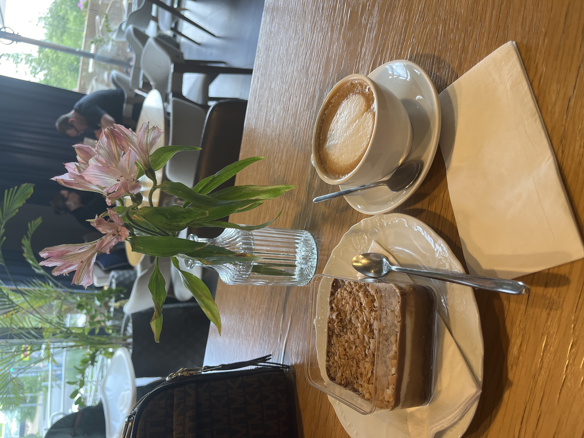 The image shows a dessert and a coffee cup on a wooden table in a cafe. The dessert is in a clear plastic container and appears to be layered with chocolate and cream, topped with crumbled nuts or pastry. A silver spoon rests on the plate beneath the dessert. Next to it is a white coffee cup with a saucer, containing a cappuccino with latte art. A small vase with pink flowers and green leaves is positioned behind the dessert and coffee. A brown patterned bag is visible in the upper left corner. In the background, blurred figures of people can be seen seated at tables, with large windows revealing an outdoor view of greenery and possibly streetlights. The overall setting suggests a modern cafe environment. This scene is likely in Kazan, Russia, based on the user's prompt.