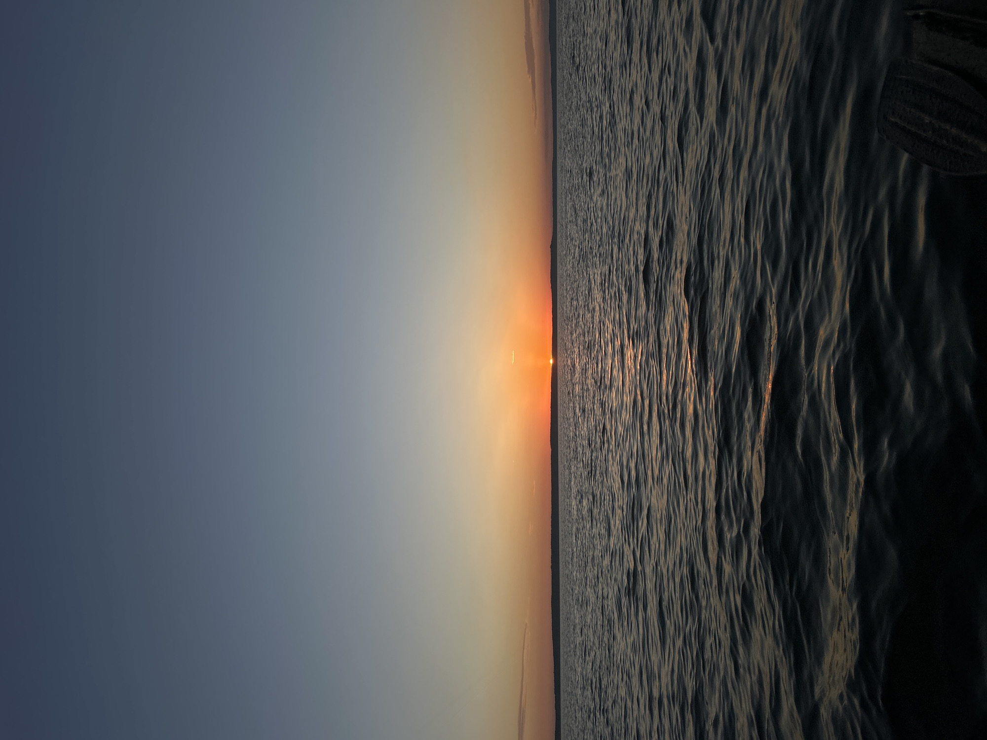 The image depicts a tranquil sunset over a body of water, likely the Kama River near Chaikovsky, Russia. The sky transitions from a pale blue at the top to a gradient of orange and yellow near the horizon, where the sun is setting. A thin, dark line of land is visible on the horizon. The water is dark and choppy, reflecting the warm colors of the sunset. In the bottom right corner, a portion of a dark-colored oar is visible, suggesting the photo was taken from a boat or kayak. There are no people or other identifiable structures in the scene.