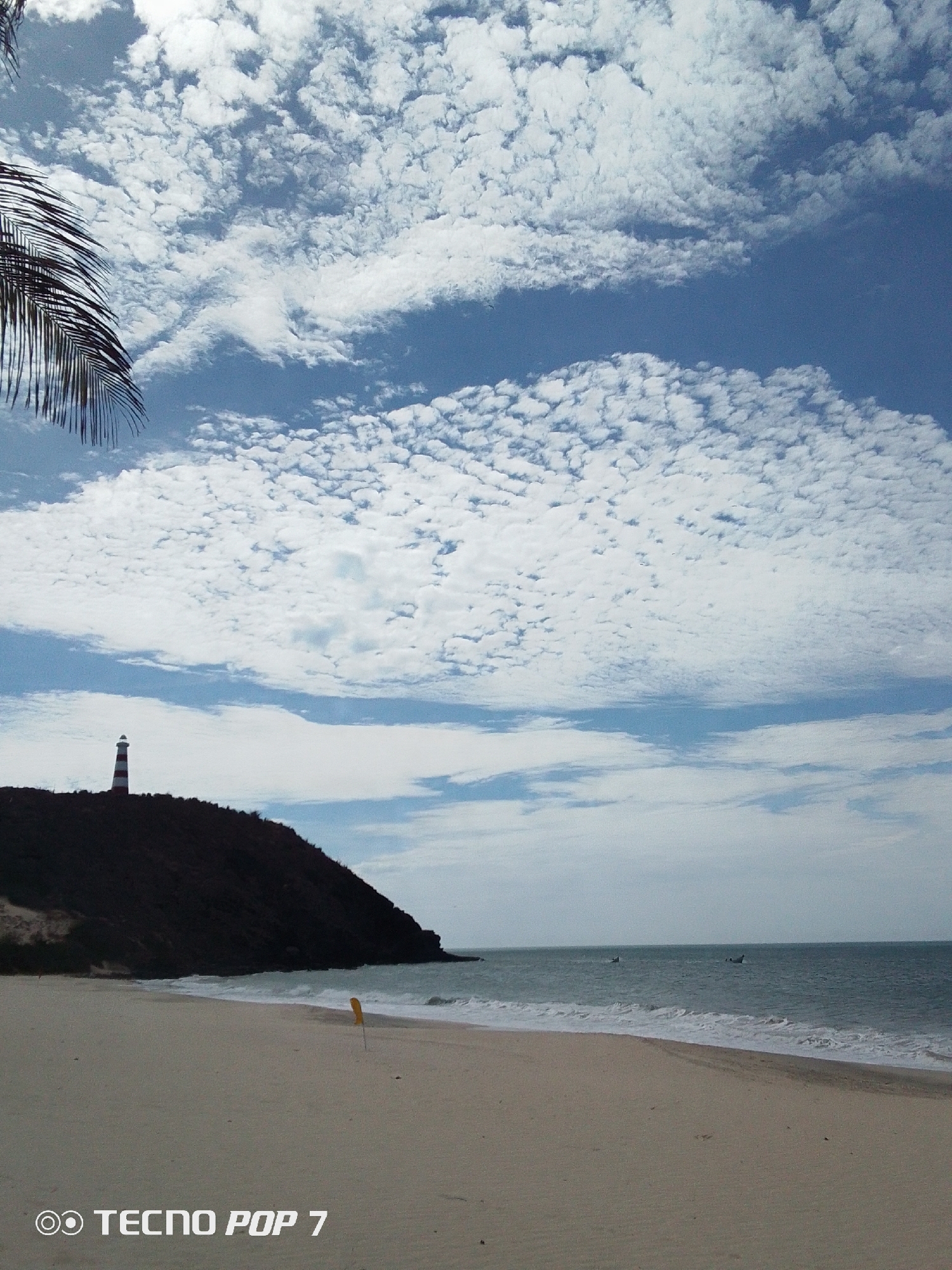 The image depicts a coastal scene in Pedro González, Venezuela. A sandy beach stretches towards the ocean. A dark, tree-covered hill rises on the left side of the image, topped with a lighthouse featuring red and white stripes. In the foreground, a yellow flag stands on the beach. The sky is blue with numerous scattered white clouds. Part of a palm tree is visible in the upper left corner. Two small boats can be seen on the water in the distance. The water's edge has light waves. A faint watermark of "TECNO POP 7" is present at the bottom of the image. There are no people visible.