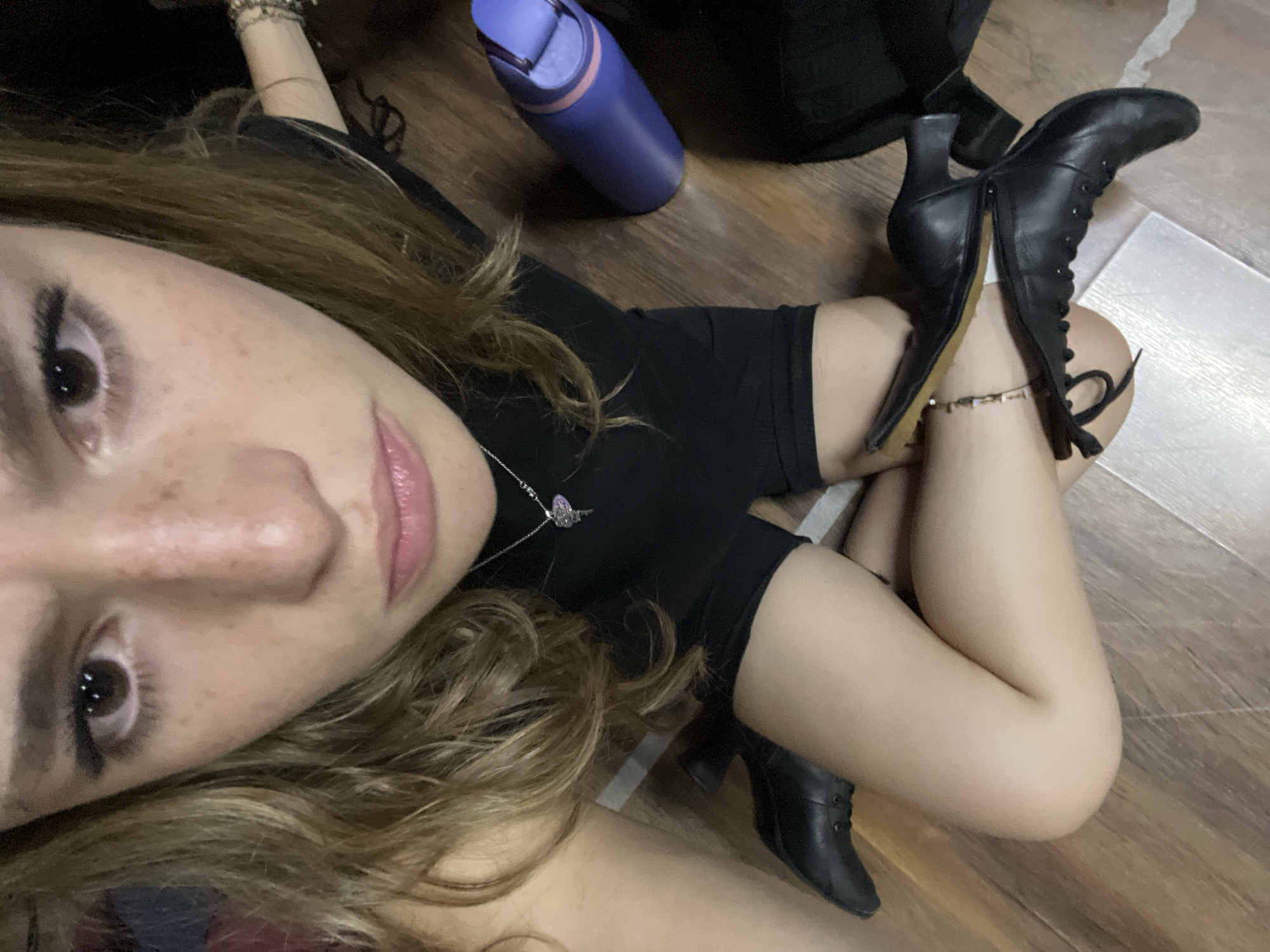 A person lies on their side on a wood floor, with a purple water bottle and a black backpack nearby. The person is wearing a black top and shorts and black heeled boots. A silver necklace is visible around their neck. The location is not discernible from the image, and the city of San Bernardino Tlaxcalancingo, Mexico cannot be confirmed.