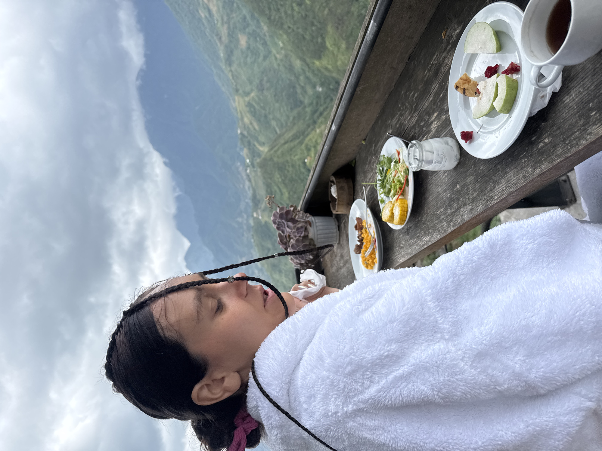 A woman with braided hair, wrapped in a white towel, is lying down on a wooden table set with food and drink. The table holds plates of food, including fruit, salad, and what appears to be corn. There is also a glass jar and a cup filled with liquid. In the background, there is a view of mountains under a cloudy sky. Based on the location of Sa Pa Ward, Vietnam, the scene suggests an outdoor dining area at a high altitude.