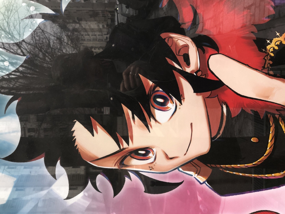 The image is a photograph of a reflective display featuring an anime-style character, taken on a street in Paris, France. The central subject is an animated character depicted from the chest up, with spiky black hair, large red-orange eyes, and light skin, exhibiting a subtle closed-mouth smile. The character has small dot-like marks on the viewer's left cheek and a stud in the left ear. Clothing consists of a dark, possibly uniform-style, garment with a gold-colored chain or trim across the chest and a visible gold button. A prominent red, ruffled or feathered element is visible on the character's right side, extending from the collar area. The character's right index finger is raised and pointed slightly upwards, partially obscuring the lower face.
Reflected in the glossy surface of the display is an urban environment consistent with Paris, France, characterized by multi-story Haussmannian-style buildings with light stone facades, bare deciduous trees, and parked cars on a street. A reflection of the photographer is also visible on the right side of the image, depicting an individual wearing a dark jacket over a lighter shirt, holding a smartphone to capture the image. A ring is visible on the photographer's right index finger, and a portion of an earbud wire is discernible near the ear.