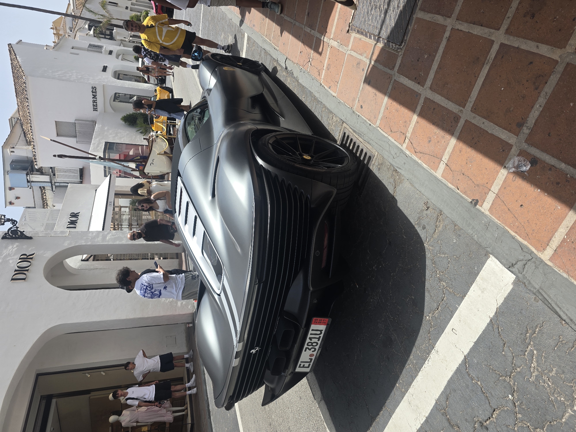An exterior, eye-level, full shot captures a matte gray Ferrari Daytona SP3 parked on the side of a street in Marbella, Spain. The luxury car, with its distinctive aerodynamic design and silver racing stripes, takes up a significant portion of the foreground. Its rear license plate, displaying "EL 381U," is clearly visible.
To the left of the car, the entrance to a Dior store is visible, featuring mannequins dressed in light-colored clothing. Several individuals are present, with two young men in white t-shirts and shorts standing outside the store. Another person, also in a white t-shirt and gray shorts, is looking down at their phone.
Further down the street, the Hermes store is identifiable by its prominent signage. Other pedestrians are scattered along the sidewalk. The street itself is paved with asphalt and bordered by a brick sidewalk. The scene suggests a sunny day in a high-end shopping district.