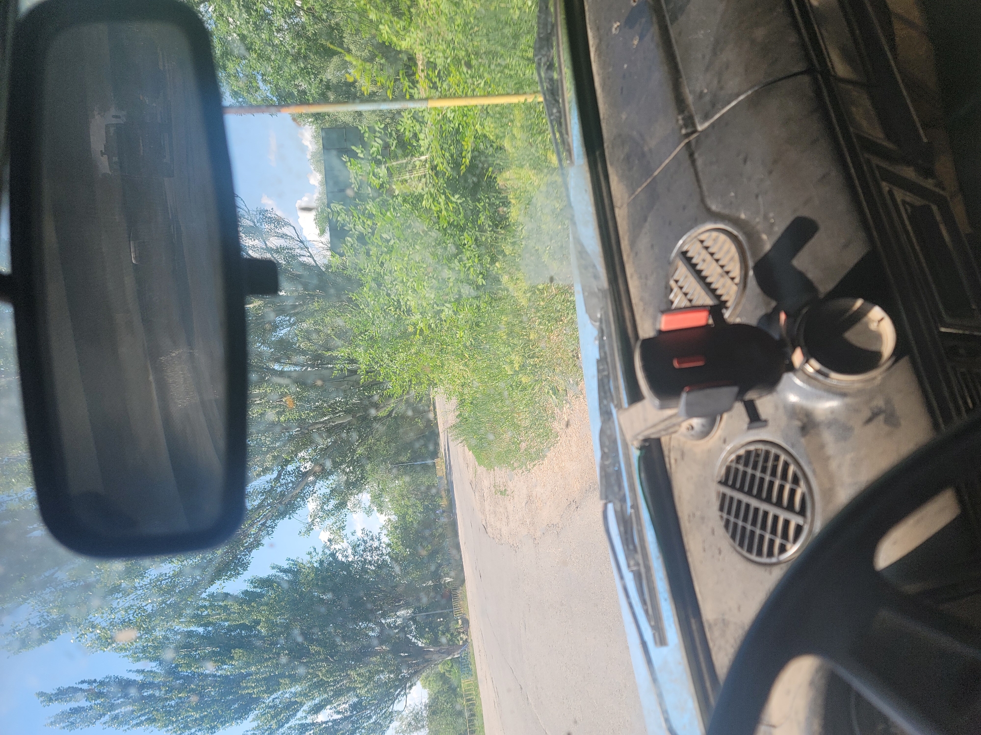 The image, taken from inside a car, shows a view through the windshield looking out at a paved road curving to the left. Tall, green trees line the road, and there is dense green vegetation on the right side. A yellow railing is visible on the left side of the road in the distance. In the foreground, the dashboard of the car is visible, with a phone mount, a circular vent, and part of the steering wheel. The rearview mirror shows a reflection of a vehicle, likely a truck, on the road behind. The sky is blue with some white clouds. The scene appears to be in a rural or suburban area. The context provided indicates the location is Pereshchepyne, Ukraine.