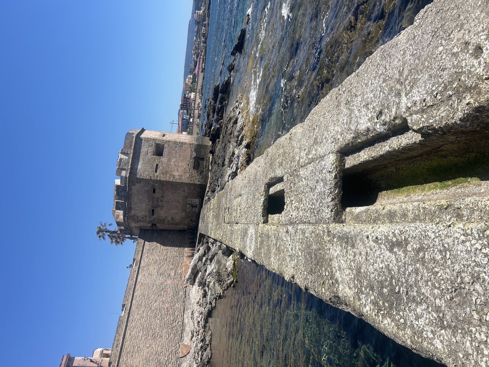 The image presents a vibrant, sunlit coastal scene in Alghero, Italy, dominated by ancient stone fortifications next to the clear Mediterranean Sea. In the immediate foreground, a broad, weathered stone structure, possibly a breakwater or walkway, extends into the water. This structure features a rough, pockmarked texture with white deposits, and has several rectangular openings or channels cut into its surface.
To the left, a robust stone fortress wall rises, constructed from large, irregular blocks. It is topped by a crenellated tower, typical of historical defensive architecture, which includes small rectangular openings or windows. A small tree or shrub can be seen growing from the top of the wall near the tower. The fortress wall continues along the shoreline, meeting a rocky coast.
The sea to the right is a striking clear blue-green, with gentle waves lapping against the natural rock formations and the base of the fortifications. In the distance, along the coastline, a settlement with buildings is visible, set against the backdrop of distant hills. The sky above is a brilliant, cloudless blue, suggesting a clear day during midday or early afternoon. The overall impression is one of historical grandeur and natural beauty under serene weather conditions. No discernible text is visible on any of the structures.