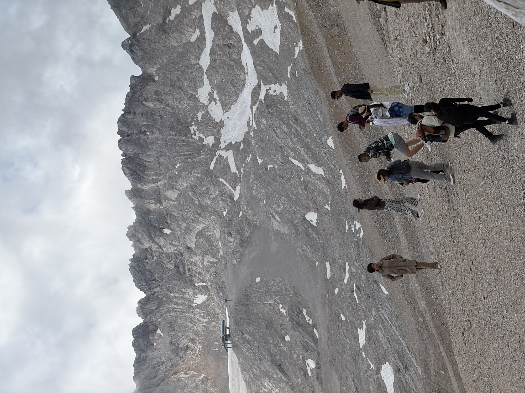The image captures a group of people on a gravelly, mountainous slope, with a dramatic rocky mountain range dominating the background. Snow patches are scattered across the mountainside. In the mid-ground, a ski lift station is visible, suggesting the location is a high-altitude area. The people, diverse in age and attire, are spread across the slope, some walking, others looking at their phones, and some interacting with each other. The lighting suggests an overcast or cloudy day. This scene is characteristic of the Zugspitze massif, the highest point in Germany, located in the municipality of Garmisch-Partenkirchen, Bavaria.