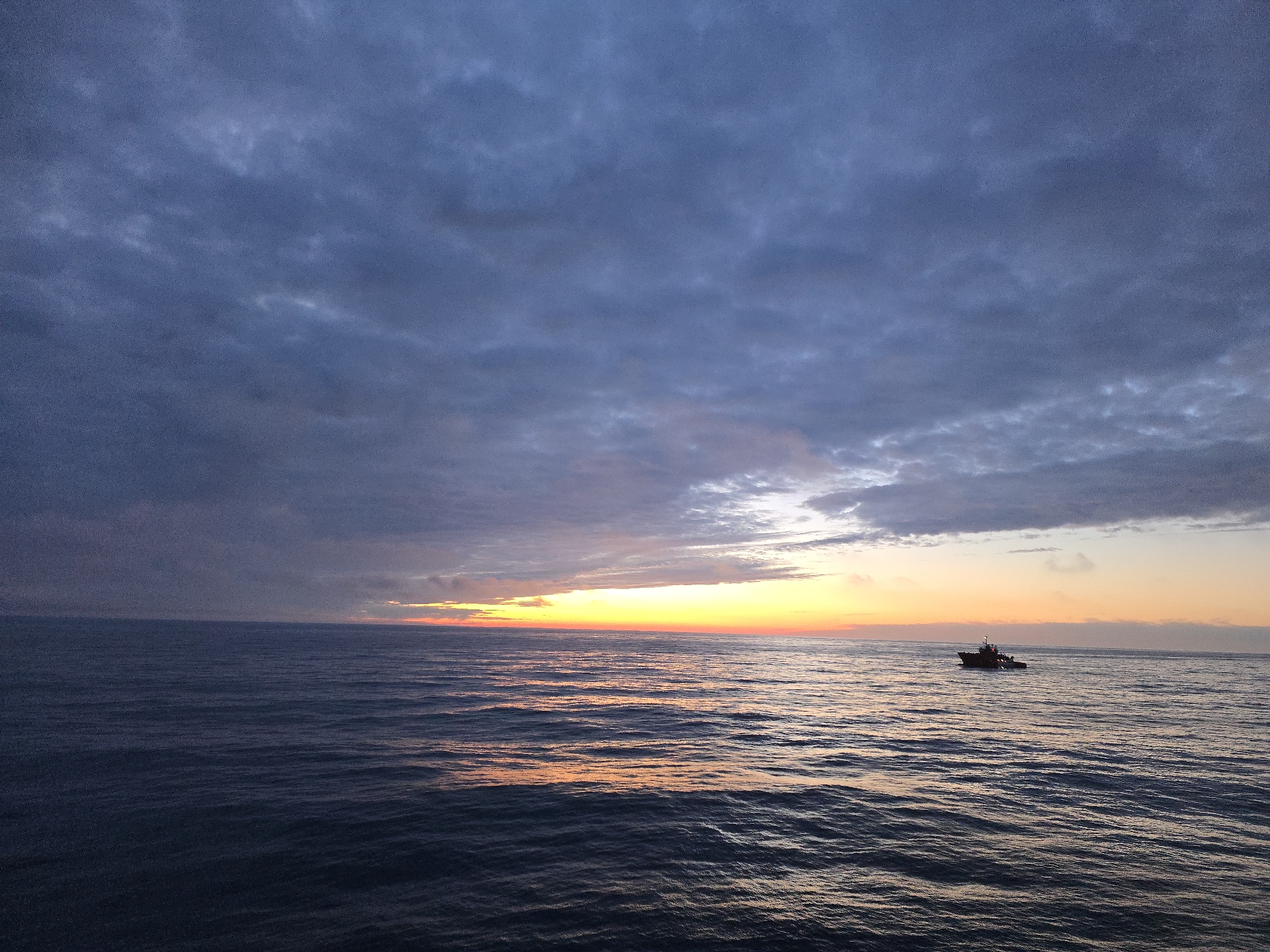 The image shows a boat on the ocean at sunset under a cloudy sky. The sky is dark blue with some lighter areas near the horizon where the sun is setting. The setting sun reflects off the water, creating a golden glow. The boat appears small and silhouetted against the light. The location is an unknown city.